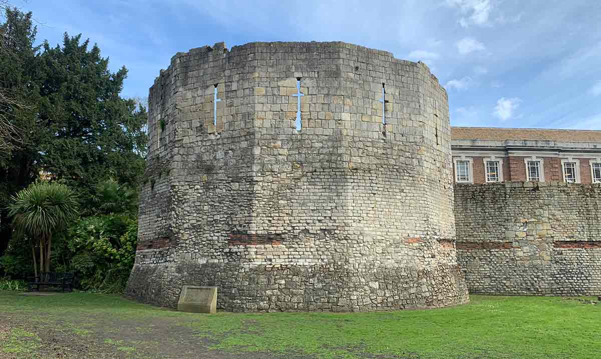 Rounded stone corner tower of the Multangular Tower in York Museum Gardens, featuring Roman brick bands, narrow vertical windows, a green lawn, and trees under a partly cloudy blue sky