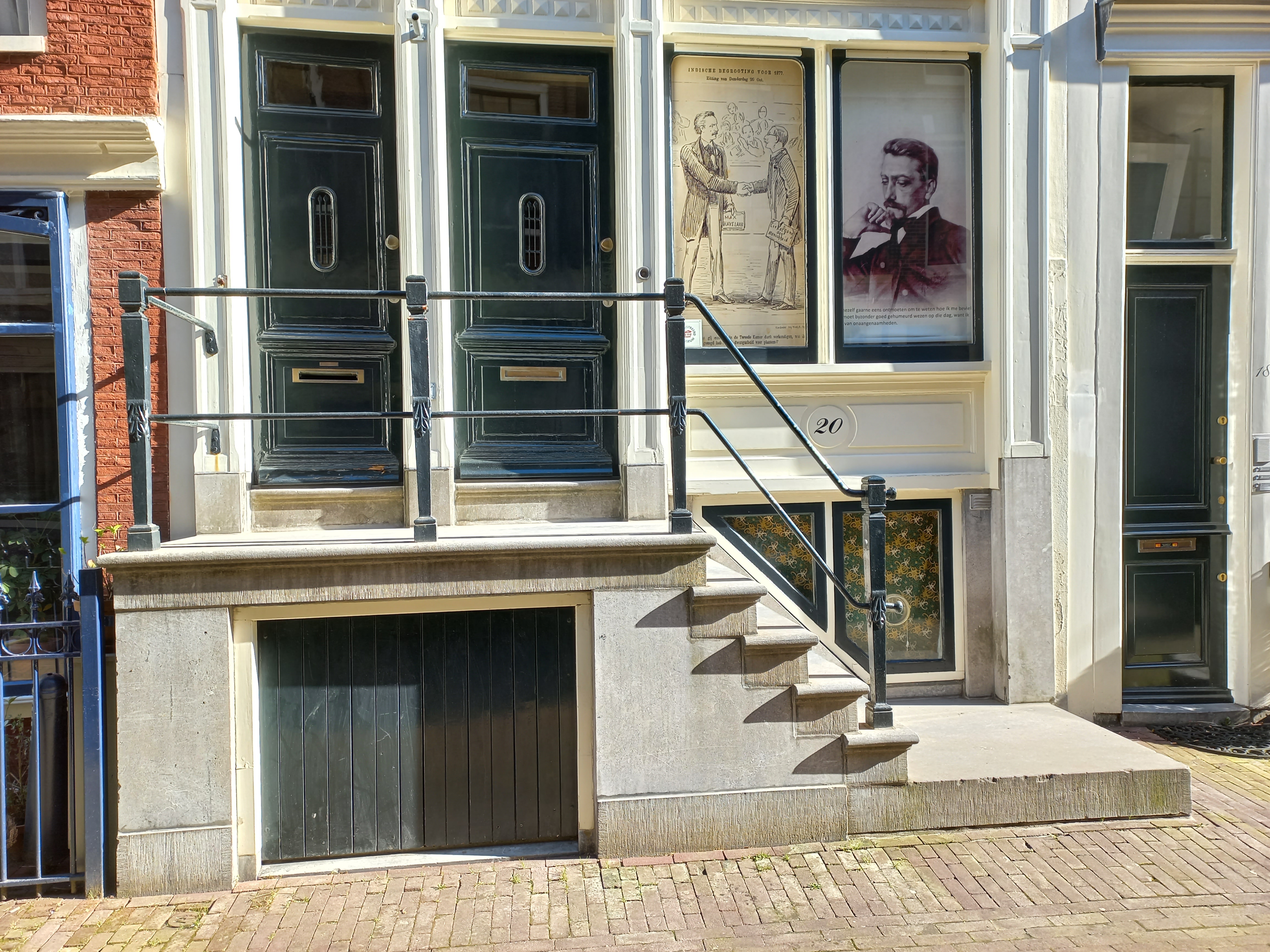 Fasada Muzeum Multatuli z dwojgiem zielonych drzwi z mosiężnymi klamkami, kamiennymi schodami i witrynami prezentującymi historyczne fotografie