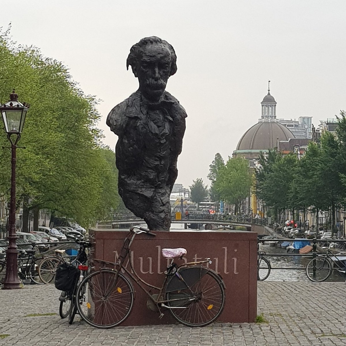 Brązowe popiersie Multatuli na cokole z widocznym napisem, zaparkowane rowery przed nim, drzewa wzdłuż ulicy i kopułowy budynek w tle.