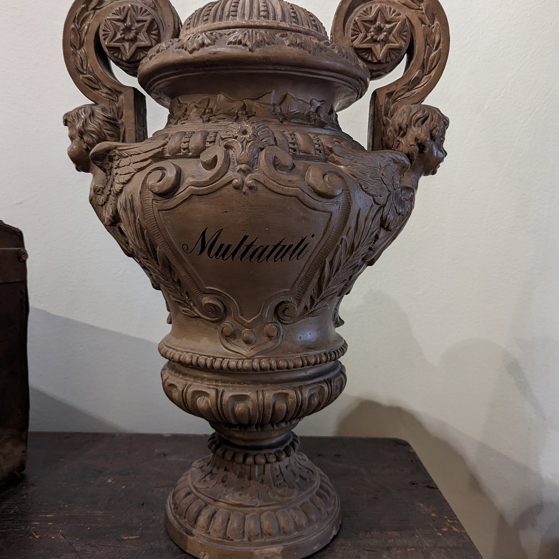 Ozdobny brązowy ceramiczny wazon z napisem 'Multatuli', misternymi rzeźbieniami i uchwytami w kształcie lwiej głowy na ciemnej powierzchni