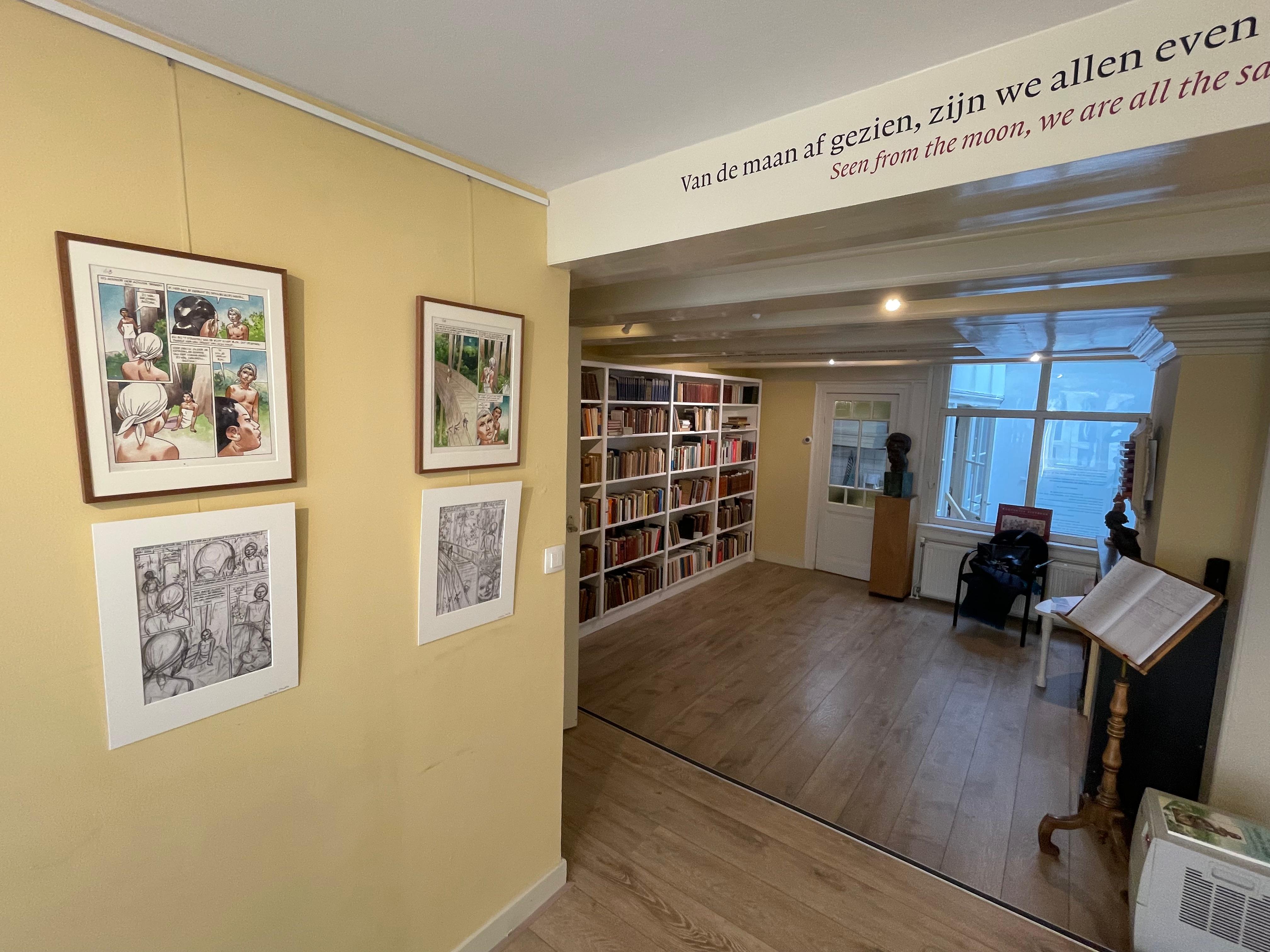 Sala muzealna z żółtymi ścianami, oprawionymi fotografiami, półkami na książki, drewnianą podłogą i wielojęzycznym cytatem na suficie