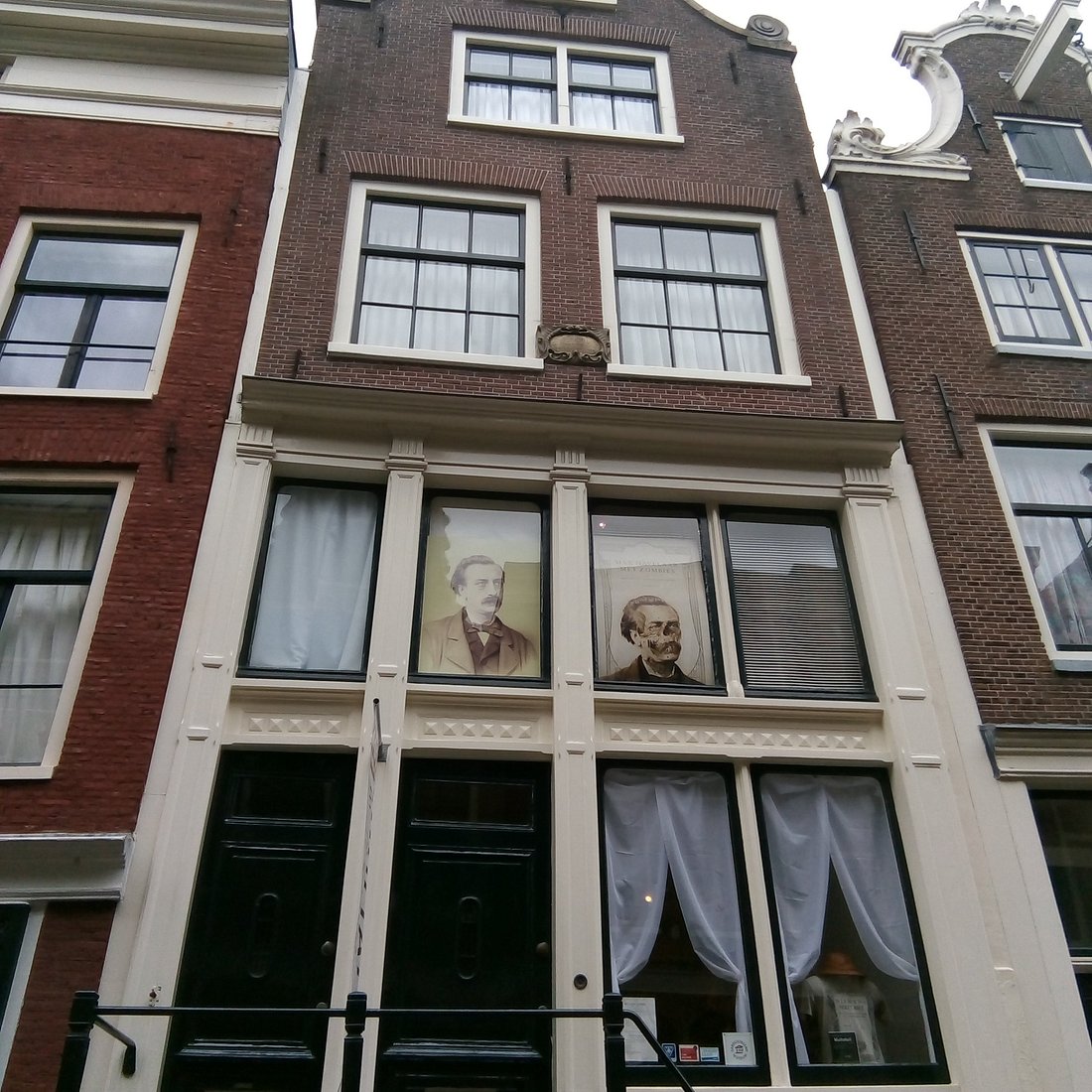 Ceglana fasada zabytkowego amsterdamskiego budynku z dużymi oknami, w których wiszą dwa portrety nad wejściem