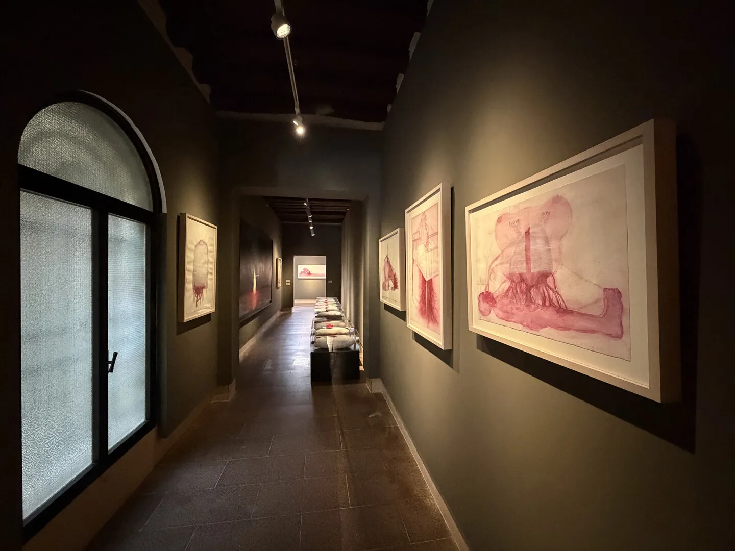 Długi korytarz galerii w Muzeum Montanelli z oprawionymi pracami w odcieniach różu oraz rzeźbami wyeksponowanymi w świetle reflektorów.