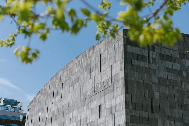 nowoczesny budynek z teksturowaną szarą kamienną fasadą i wygrawerowanym na ścianie napisem 'mumok', ujęty w ramy zielonych gałęzi drzew na tle niebieskiego nieba