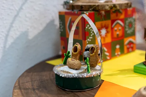 Een kleine kerststal-diorama met twee figuren, groene cactussen en een zilveren boog, geplaatst op een groene sokkel met een kleurrijke, patroonachtige achtergrond.