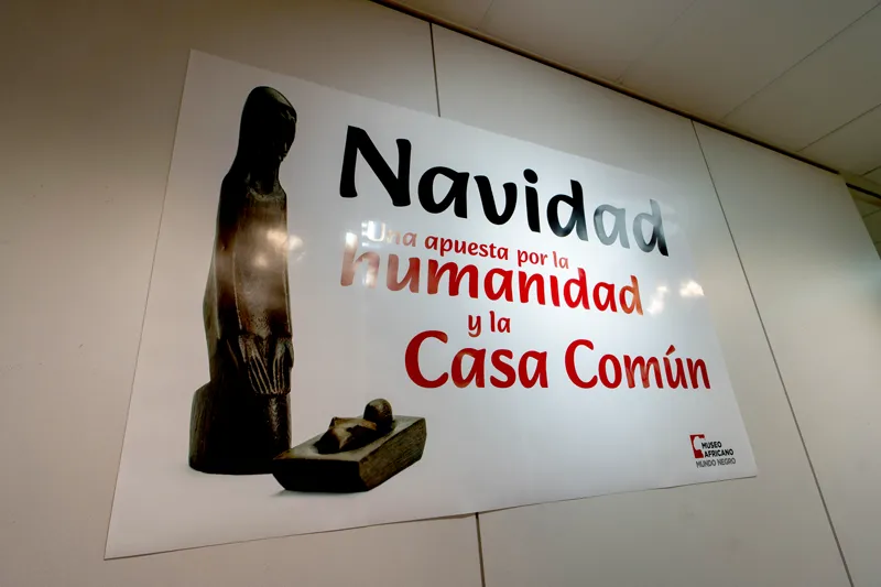 A poster on a wall showing a dark sculpture of a standing figure next to a smaller object, with text in Spanish that includes 'Navidad' and 'Casa Común'.