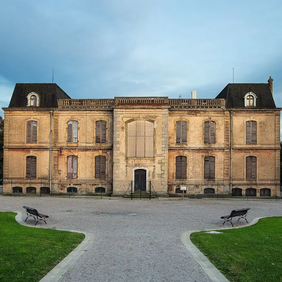 Front facade of a French Second Empire-style stone palace with a symmetrical design, two mansard-roofed pavilions, shuttered windows, and a central entrance, viewed from a gravel courtyard with two benches and green lawns
