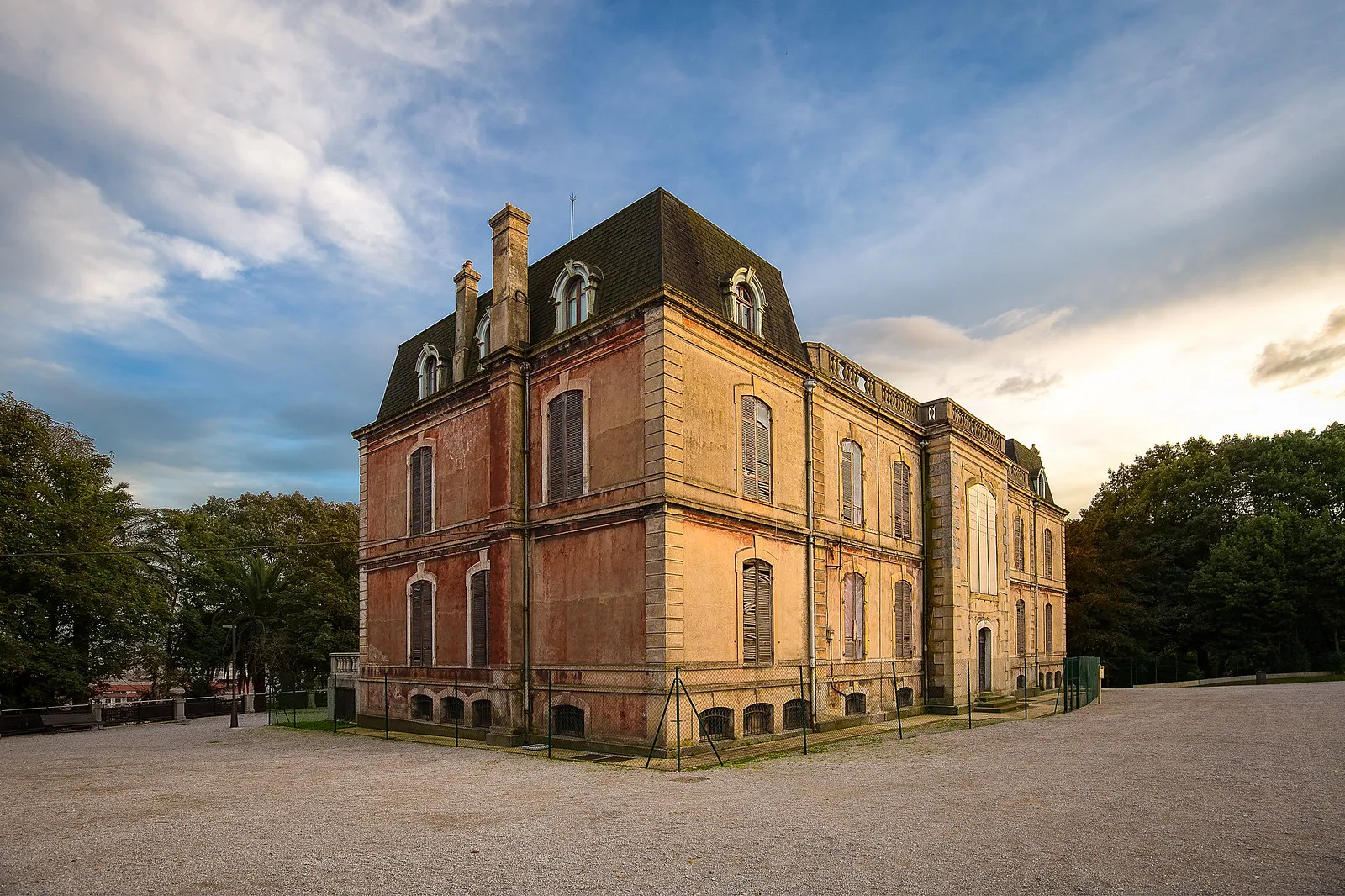 Exterior view of the Munoa Estate palace, a French Second Empire-style brick building with a dark mansard roof, dormer windows, and surrounding trees under a cloudy sky