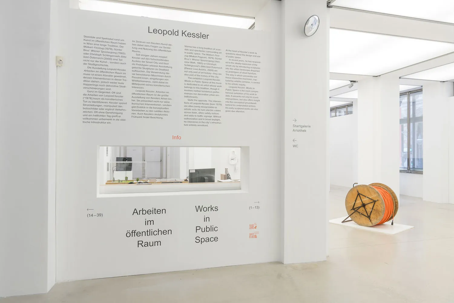 witte galerijmuur met tekstpanelen en een vitrine met een kantooropstelling, met een groot oranje en zwart cilindrisch kunstwerk op de vloer