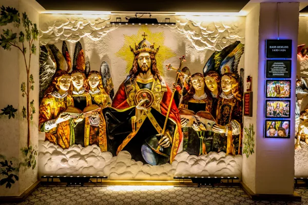 Groot verlicht gipsreliëf van een gekroonde centrale figuur geflankeerd door engelenmuzikanten tussen wolken, tentoongesteld op een museummuur met een Musea Sculpta informatiepaneel aan de rechterkant