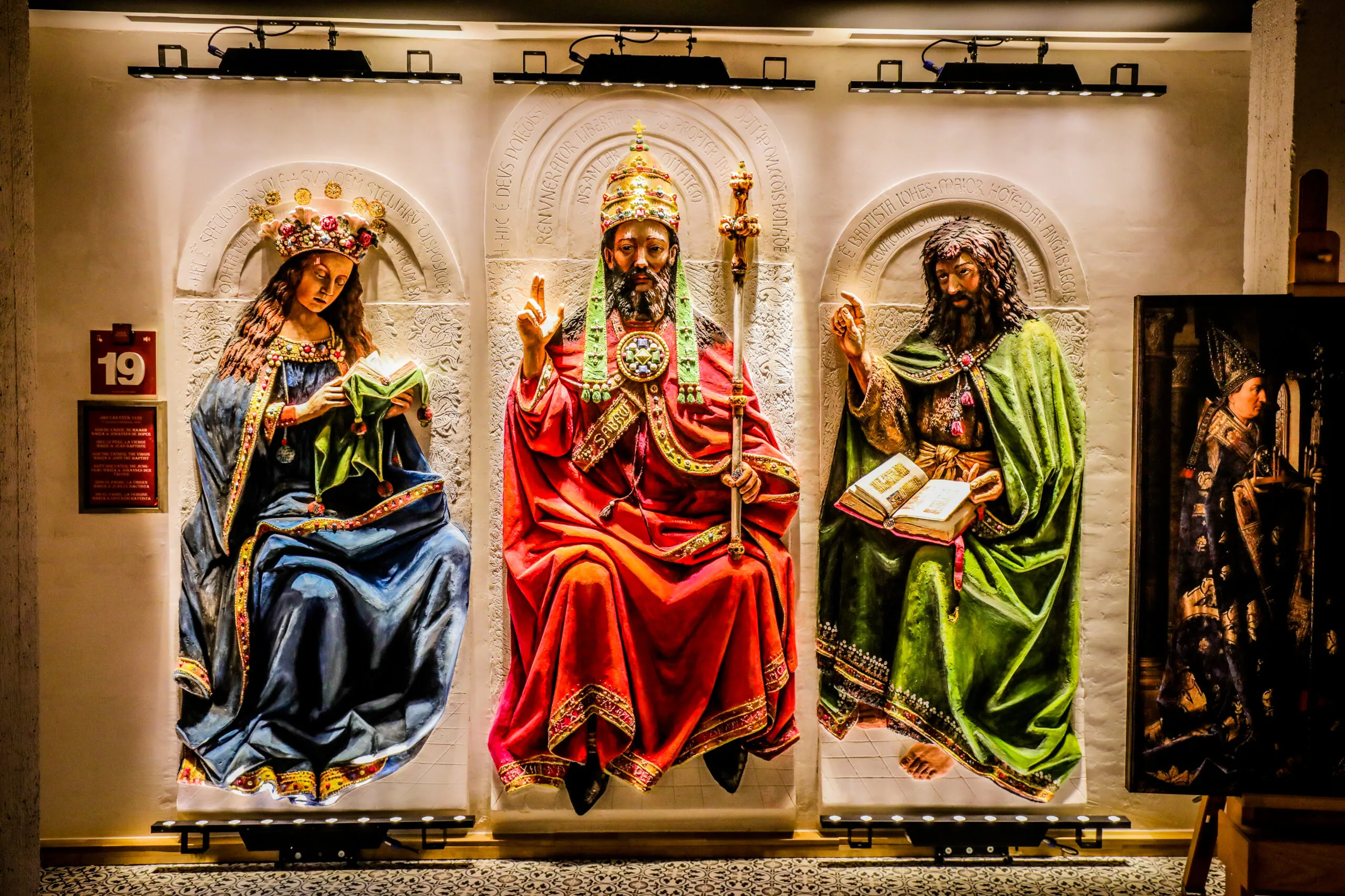 Tres grandes paneles de relieve en yeso que representan figuras religiosas con túnicas en azul, rojo y verde, expuestos en una pared de la galería con iluminación de riel arriba y una placa numerada con el 19 a la izquierda