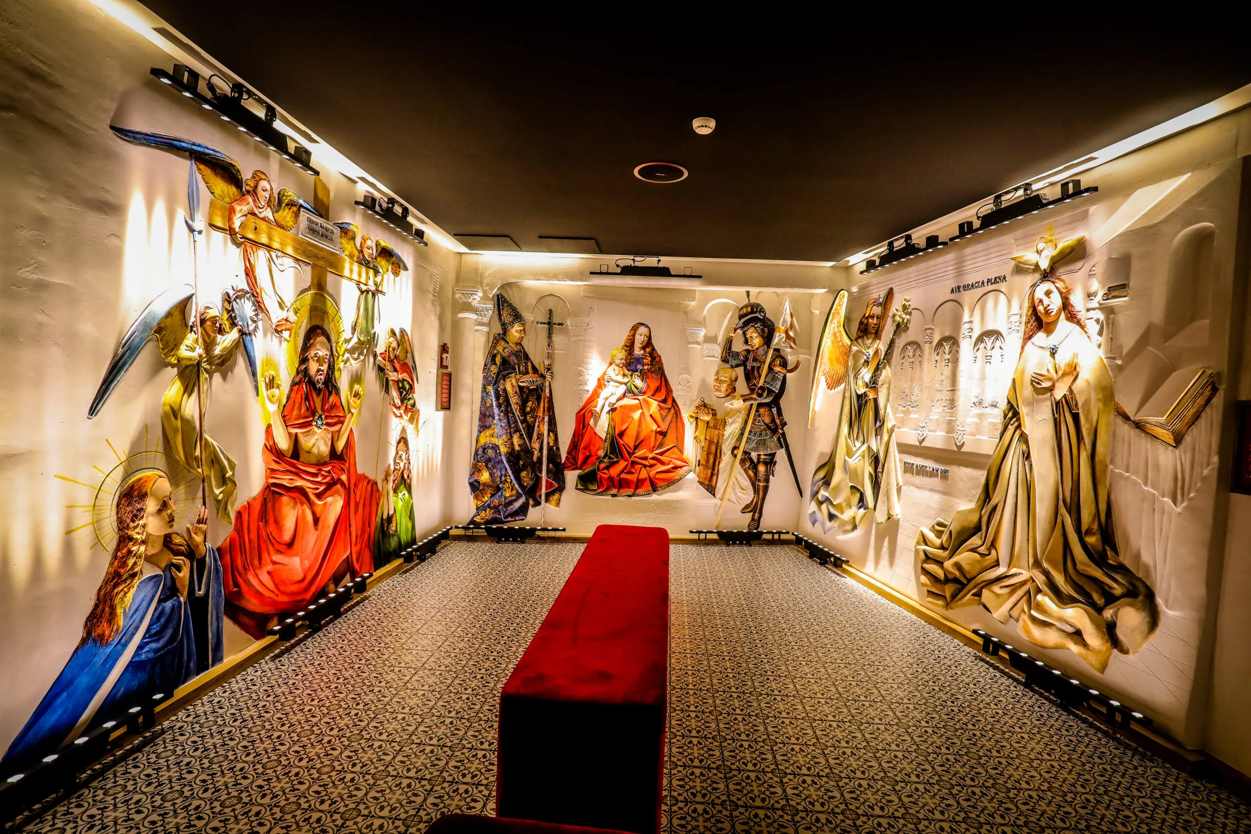 Sala de exposición interior con relieves de yeso a gran escala que representan figuras religiosas en las paredes, con un banco rojo en el centro