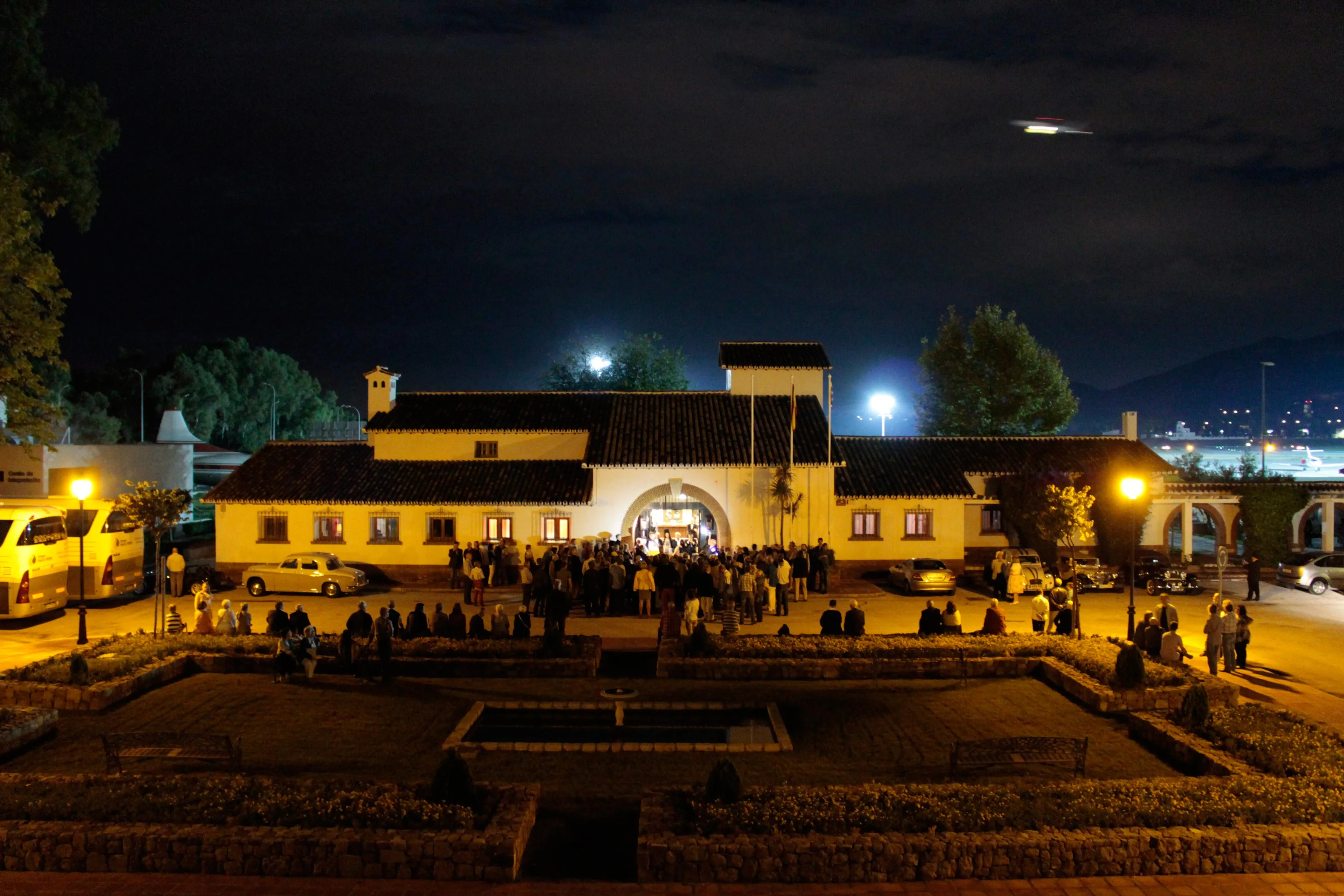 Vista nocturna del iluminado edificio histórico blanco de la terminal del Museo Aeronáutico de Málaga con una multitud reunida frente a la entrada arqueada y un jardín con setos en primer plano bajo un cielo oscuro con una luna visible.