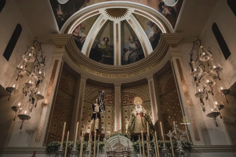 Interior ornamentado de un recinto religioso que presenta una cúpula pintada, grandes lámparas de araña y dos tronos procesionales rodeados de velas.
