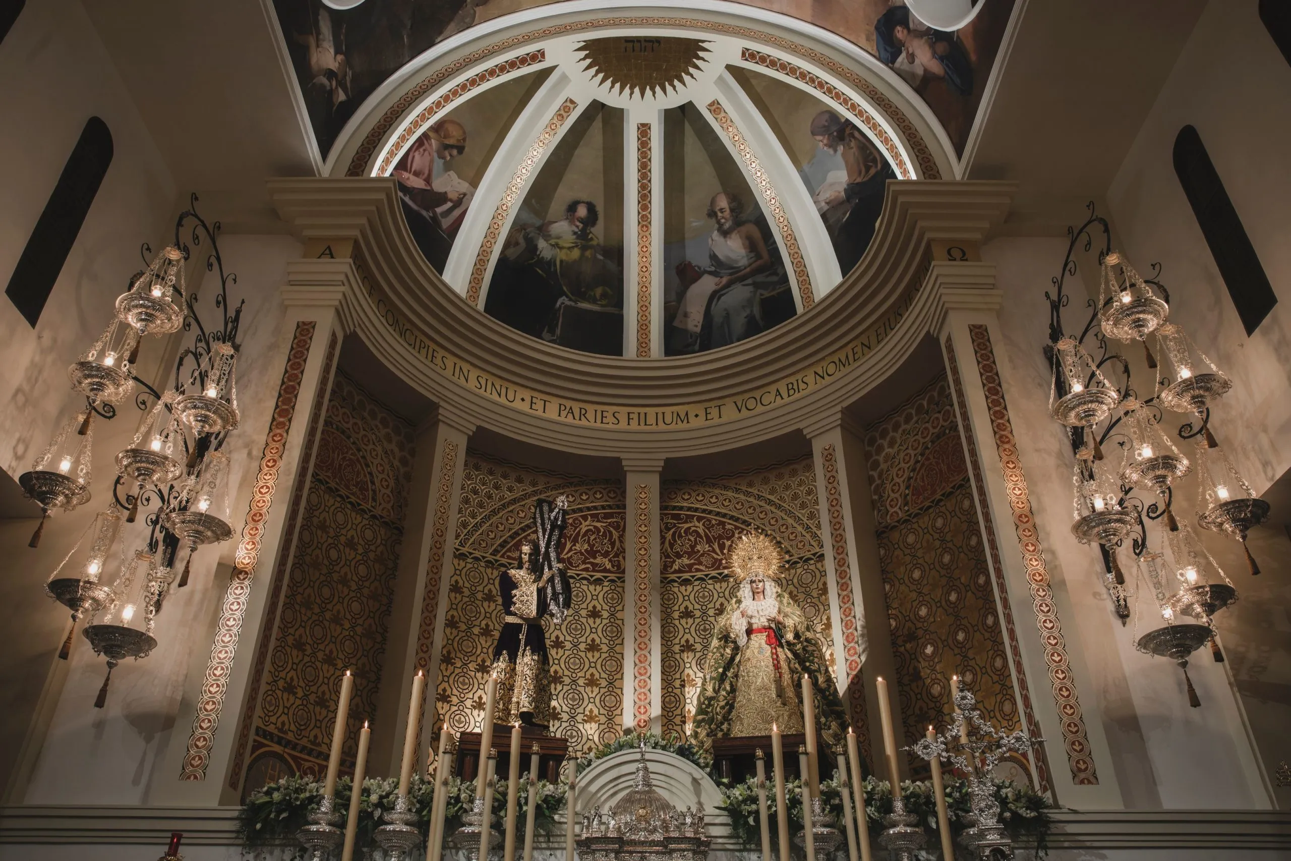Interior de iglesia ornamentado con un techo abovedado con figuras pintadas, grandes lámparas de araña de cristal y un altar con una estatua de la Virgen María coronada y un crucifijo