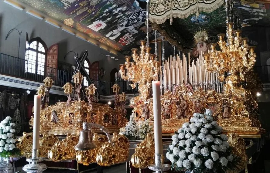 Tronos procesionales dorados y ornamentados con velas blancas altas y arreglos florales blancos dentro de una sala de museo con techo pintado y ventanas arqueadas