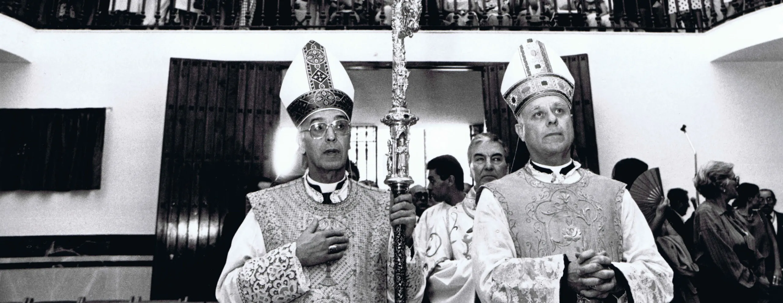 Dos clérigos que llevan mitras y ornamentos ornamentados están de pie uno al lado del otro sosteniendo un bastón ceremonial alto en una fotografía en blanco y negro.