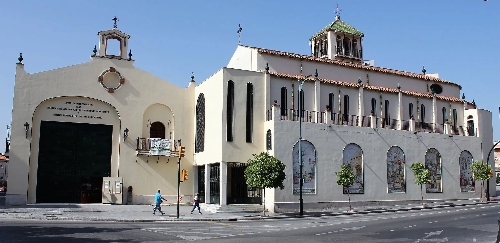 Complejo de edificios blancos con ventanas arqueadas, campanario y una gran puerta de entrada arqueada bajo un cielo azul despejado con peatones en la acera
