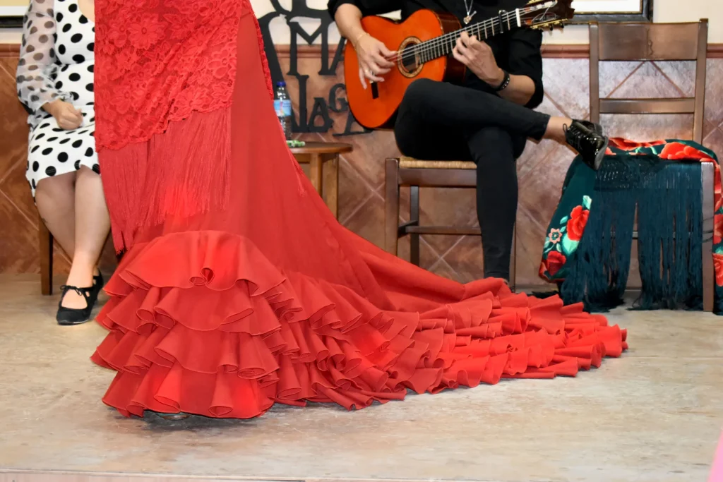 Un largo vestido de flamenca rojo con una cola de volantes extendida por el suelo, un guitarrista tocando la guitarra clásica de fondo y un mantón de flecos verde azulado drapeado sobre una silla