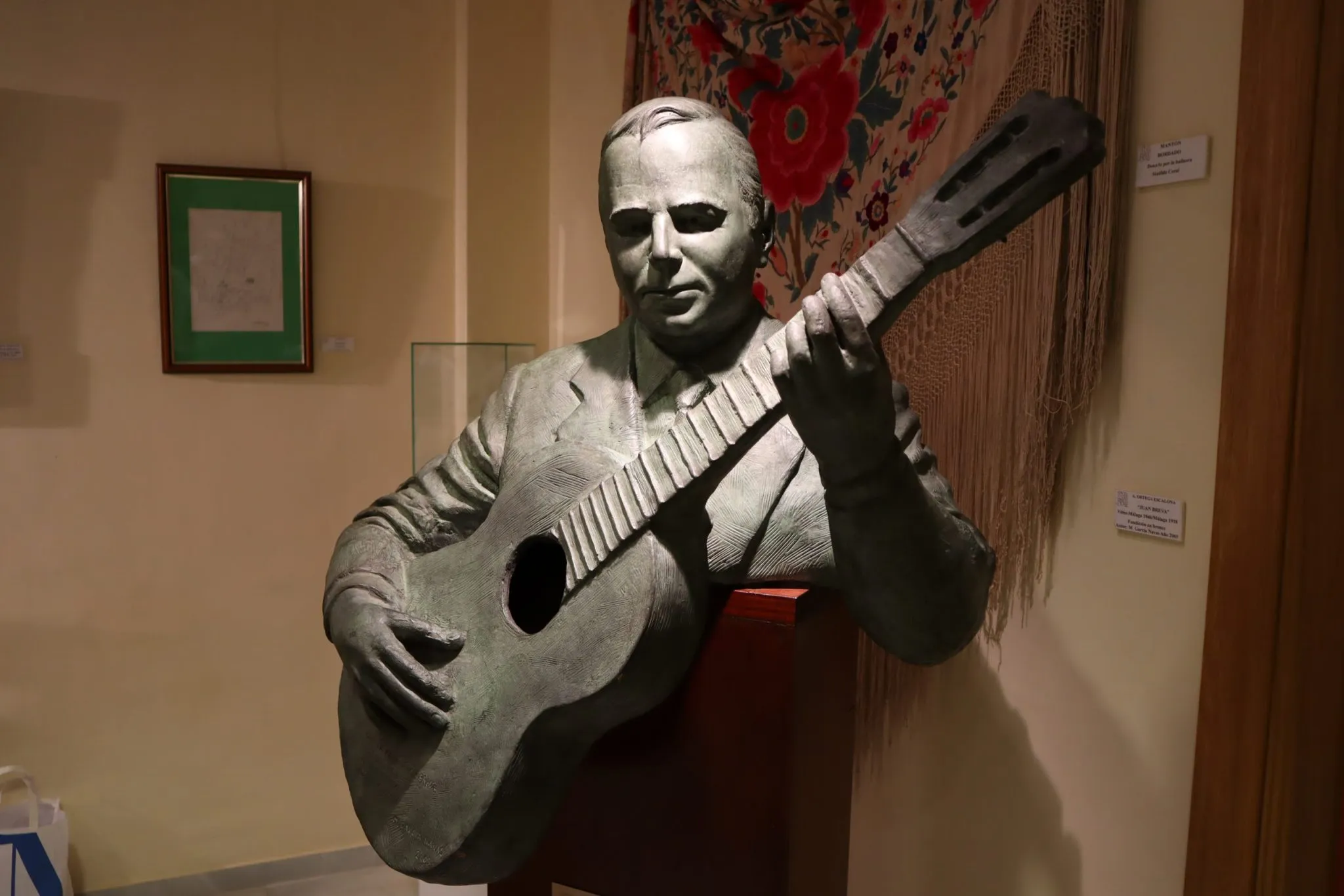 Escultura de bronce de un hombre sentado tocando una guitarra clásica, expuesta en una galería de museo con obras de arte enmarcadas y un tejido decorativo al fondo