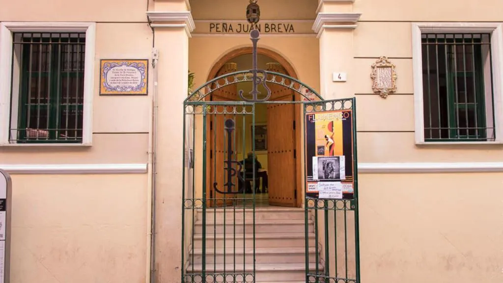 Vista exterior de la entrada a la Peña Juan Breva, que presenta una fachada de color crema, una puerta en arco con una verja metálica y el nombre del lugar inscrito sobre el arco.