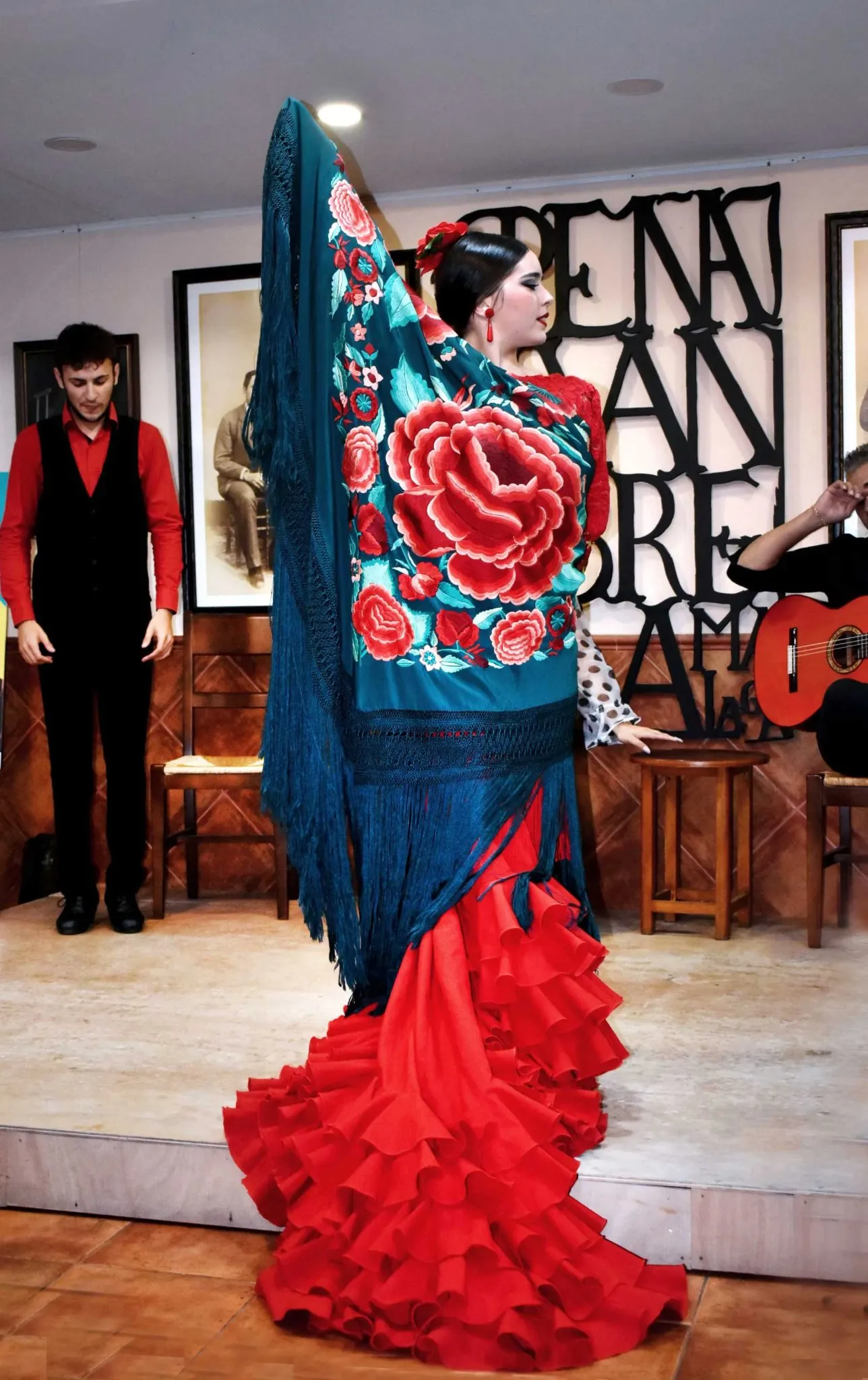 Una bailaora de flamenco con un vestido rojo de volantes y un mantón azul de flores actúa en el escenario junto a un guitarrista y un hombre que permanece al fondo, frente a un cartel en la pared que dice 'PEÑA JUAN BREVA'.