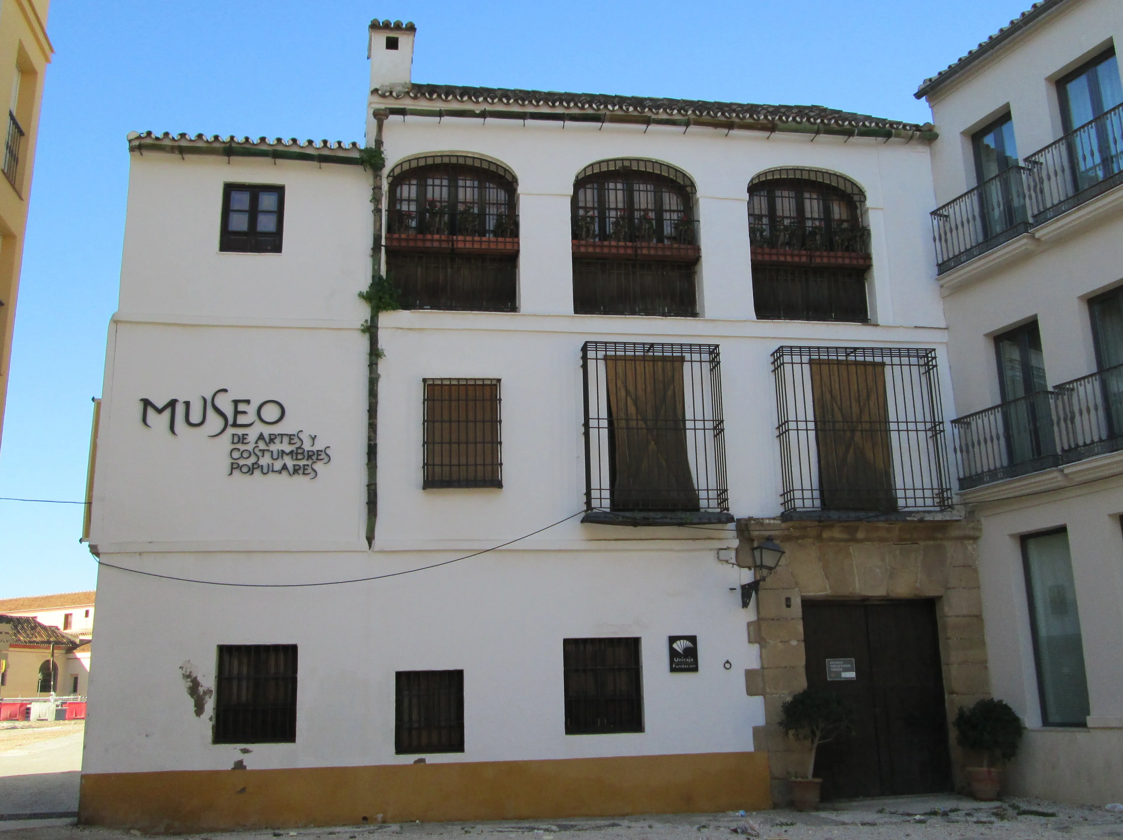 Fachada blanca de un edificio de tres plantas con 'MUSEO DE ARTES Y COSTUMBRES POPULARES' pintado en un lateral, caracterizada por ventanas arqueadas con rejas de hierro y balcones