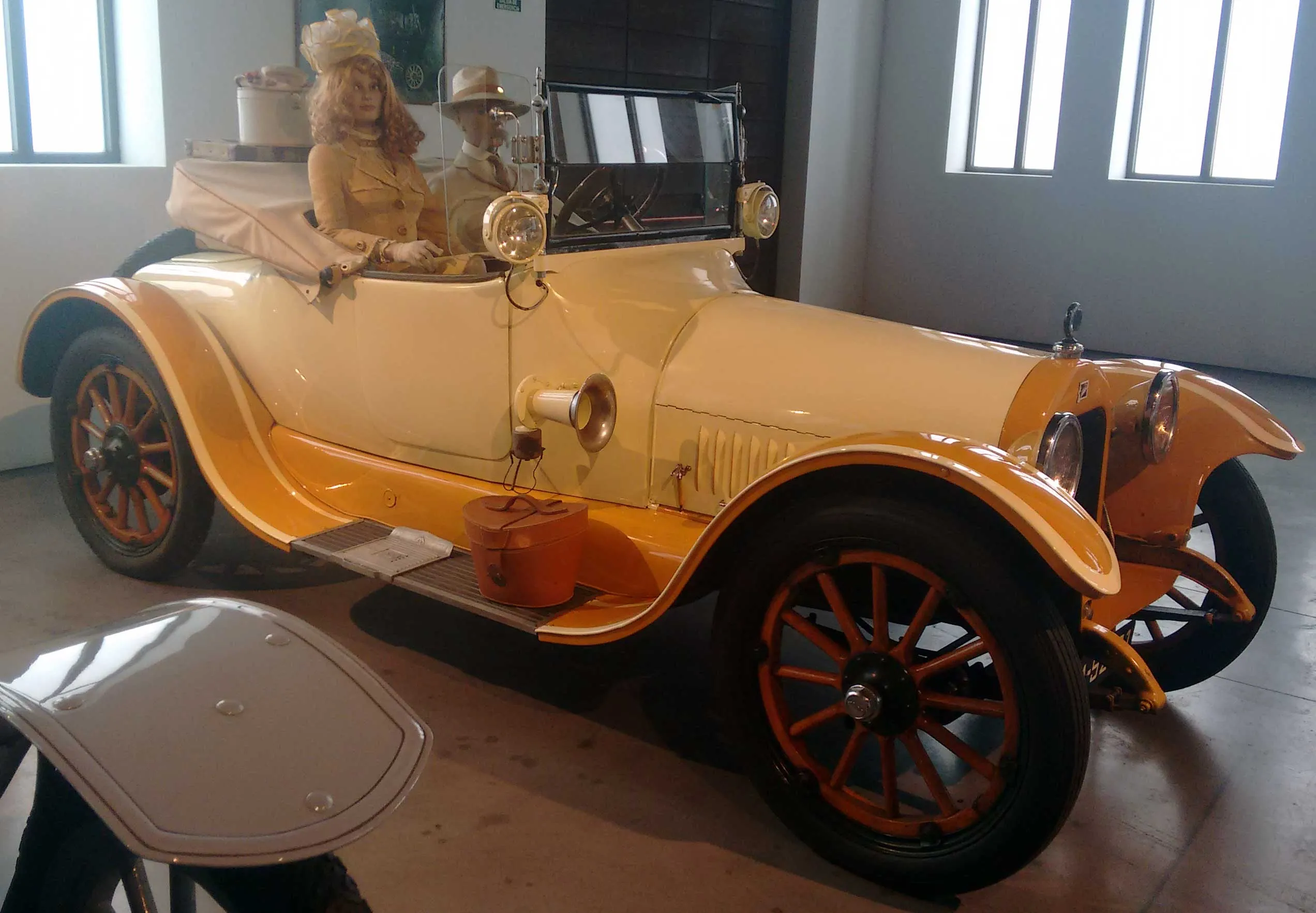 Automóvil Buick de 1916 en color crema y naranja, expuesto en una galería de museo con maniquíes vestidos de época sentados en su interior.