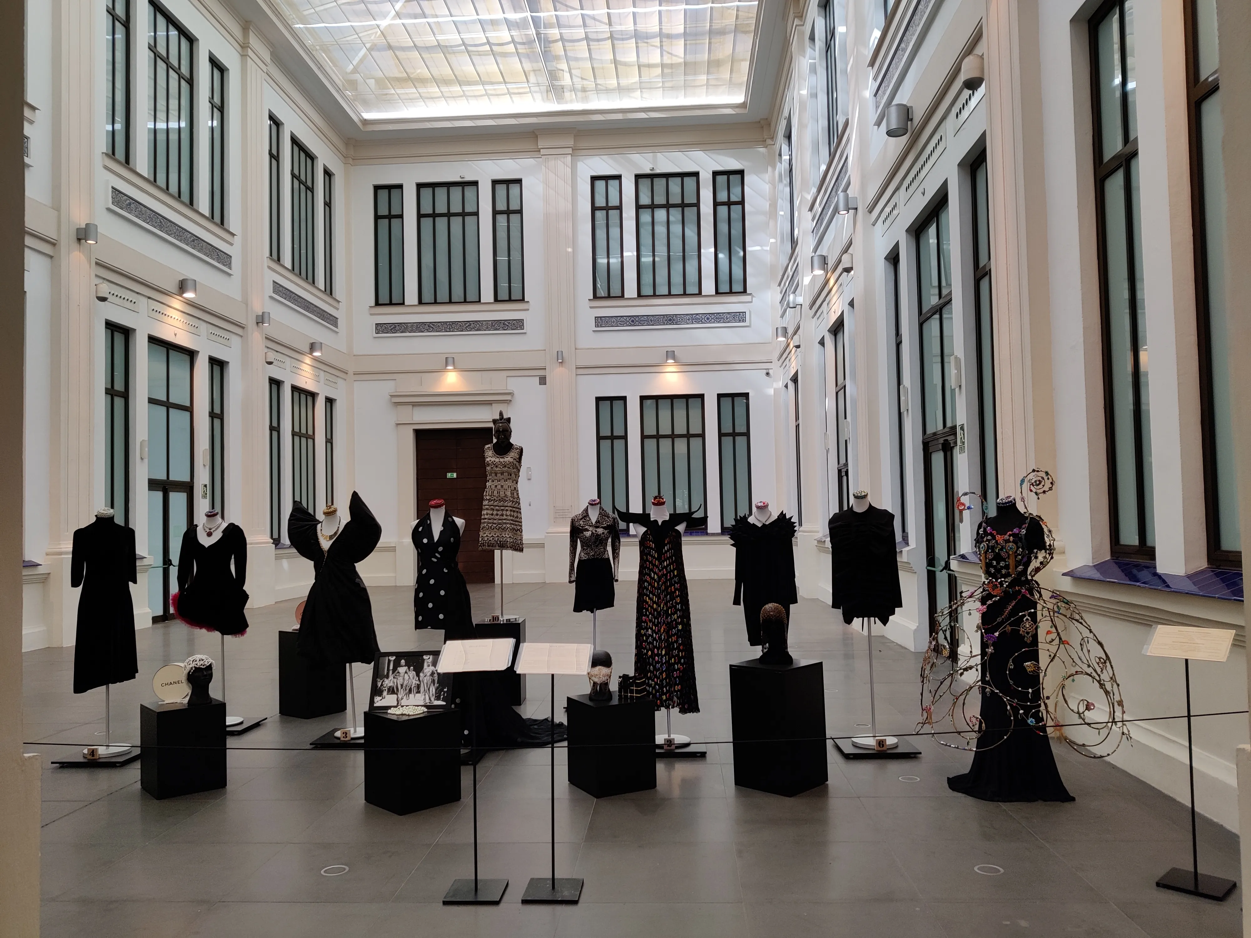 Una gran sala de museo con techos altos, una claraboya de cristal y ventanales, llena de maniquíes con prendas de moda negras sobre pedestales, con una escultura decorativa de una bicicleta en el lado derecho.