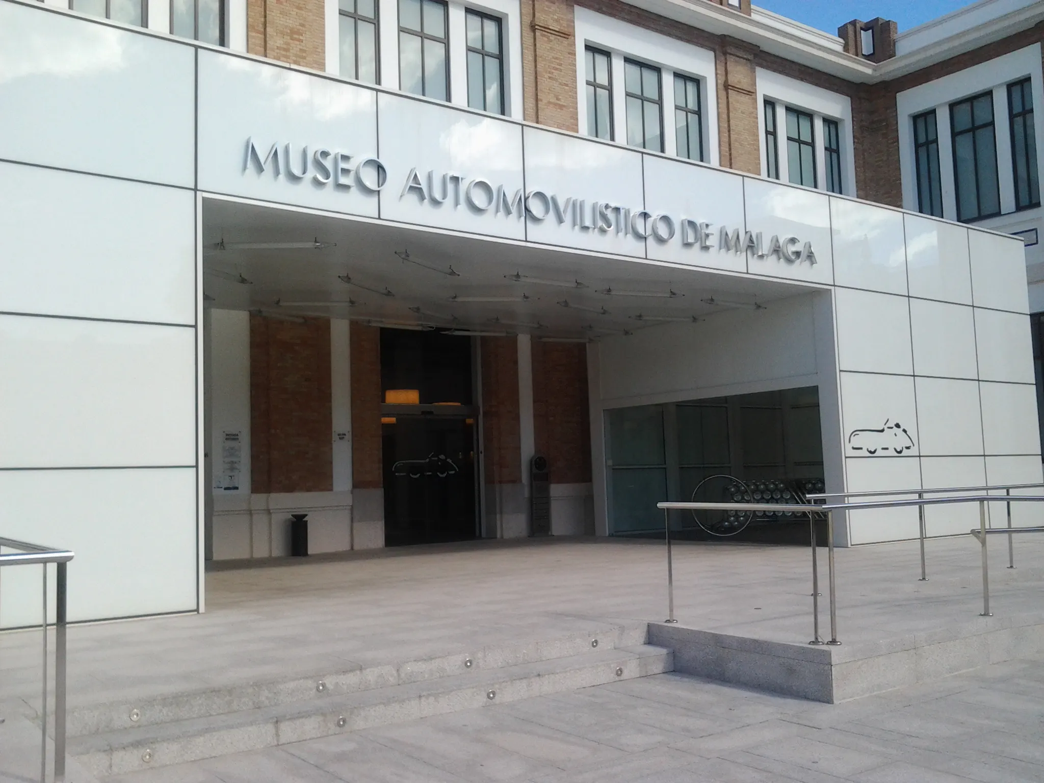 Vista exterior de la entrada del Museo Automovilístico de Málaga con señalización grande en blanco y escalones que conducen a las puertas de cristal.