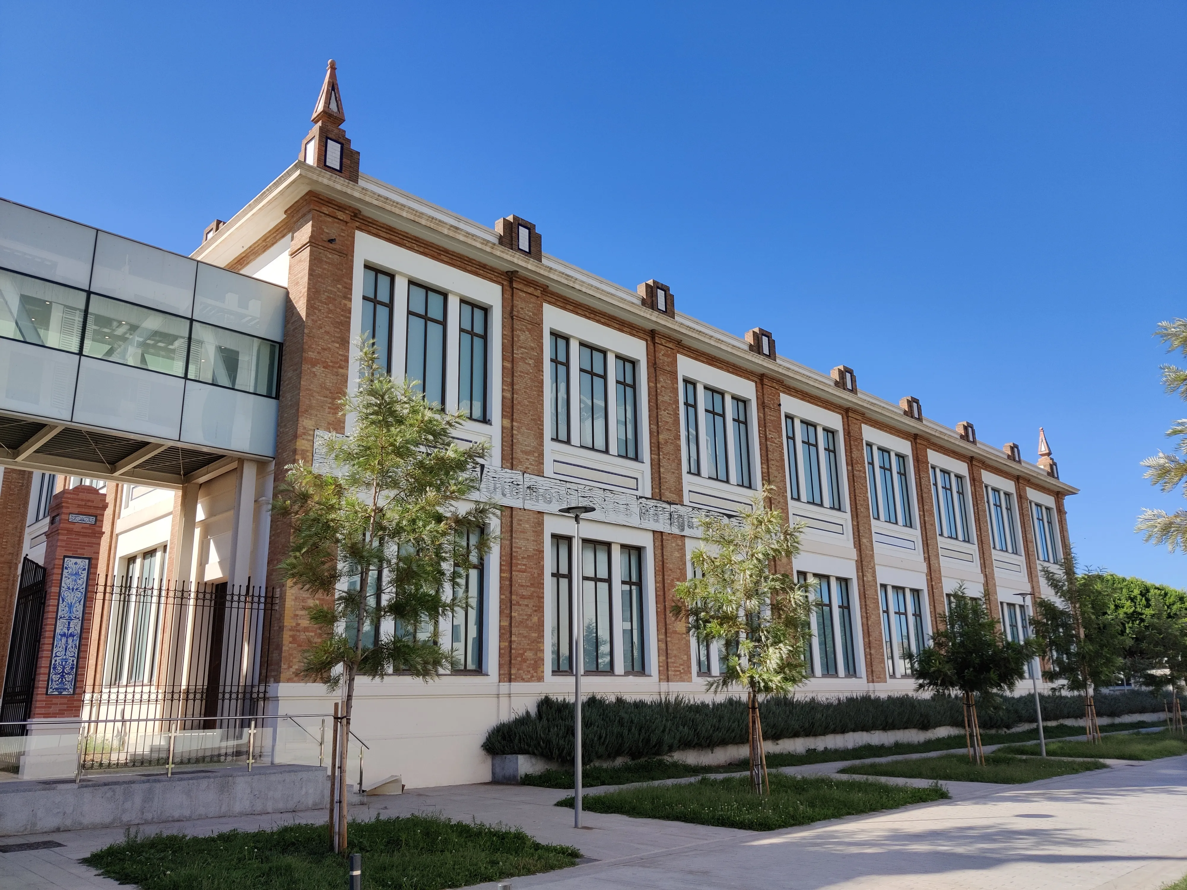Exterior del Museo del Automóvil y la Moda de Málaga, un gran edificio de ladrillo con detalles en blanco y una moderna extensión de cristal bajo un cielo azul.