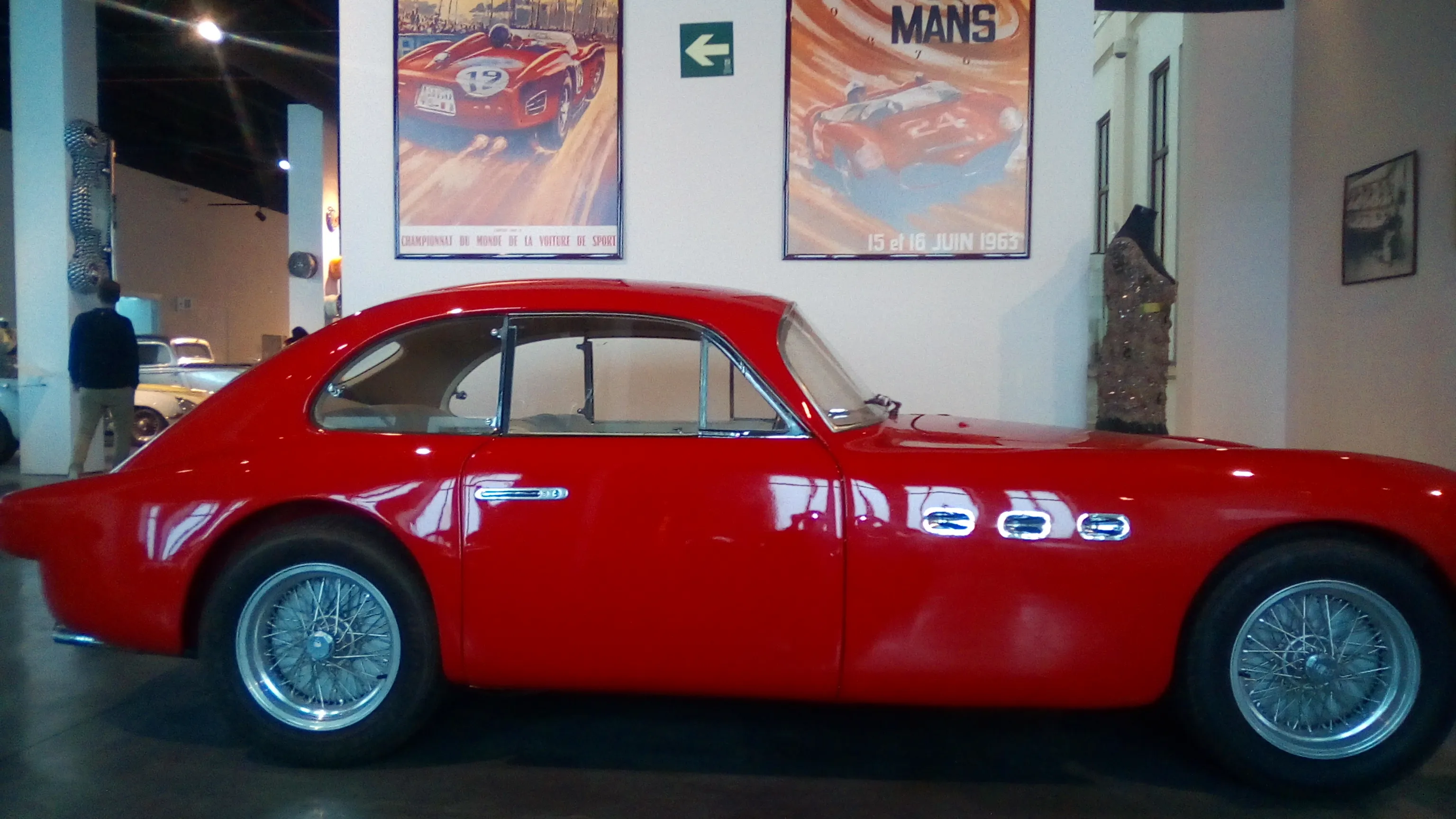 Un cupé Maserati clásico de color rojo expuesto en un museo, con pósteres de carreras de Le Mans en la pared detrás