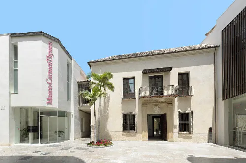 Patio soleado del Museo Carmen Thyssen Málaga con una palmera, una fachada histórica blanca con balcones de hierro y una entrada moderna de cristal a la izquierda que lleva el nombre del museo en letras rojas.