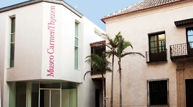 Vista exterior del Museo Carmen Thyssen Málaga, que muestra un edificio moderno de color blanco con el nombre del museo en letras rojas junto a un edificio tradicional con balcones y una palmera.