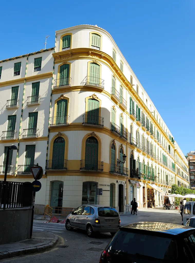 Edificio esquinero blanco con detalles amarillos y contraventanas verdes, el Museo Casa Natal Picasso en la Plaza de la Merced de Málaga bajo un cielo azul despejado