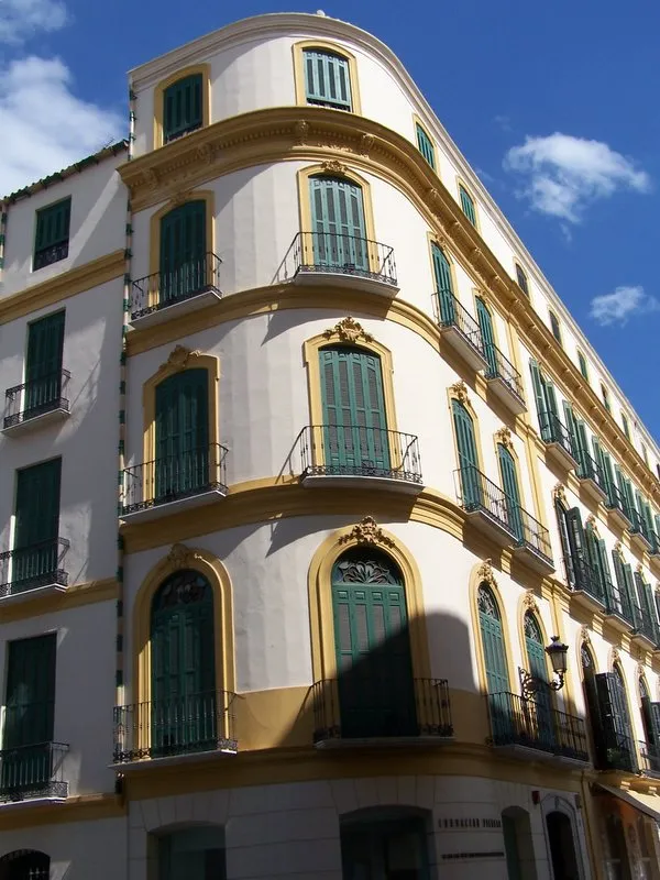 Edificio esquinero blanco y amarillo con contraventanas verdes y balcones de hierro forjado bajo un cielo azul