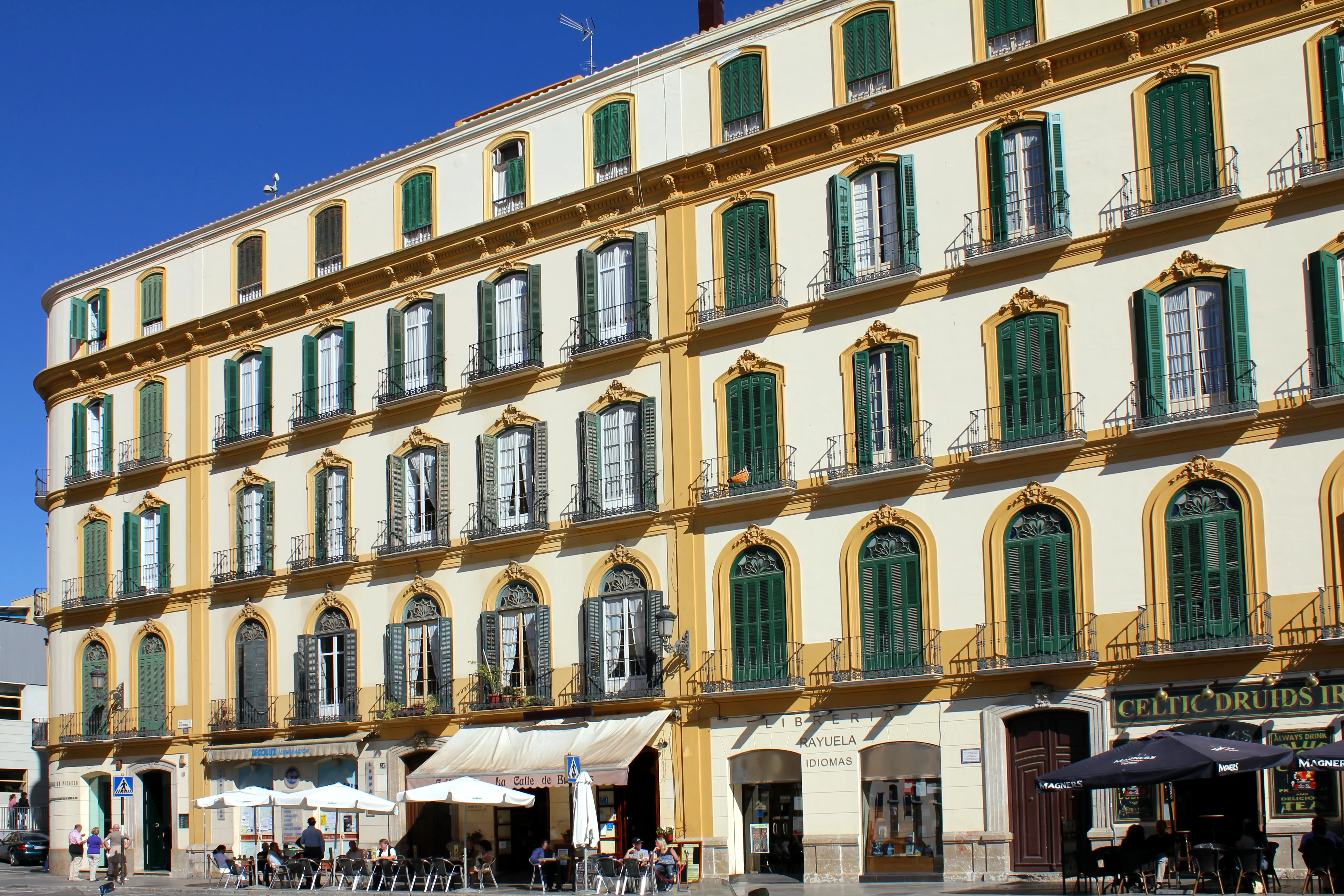 Edificio amarillo de varias plantas con contraventanas verdes y ventanas arqueadas en una plaza soleada con terrazas al nivel de la calle
