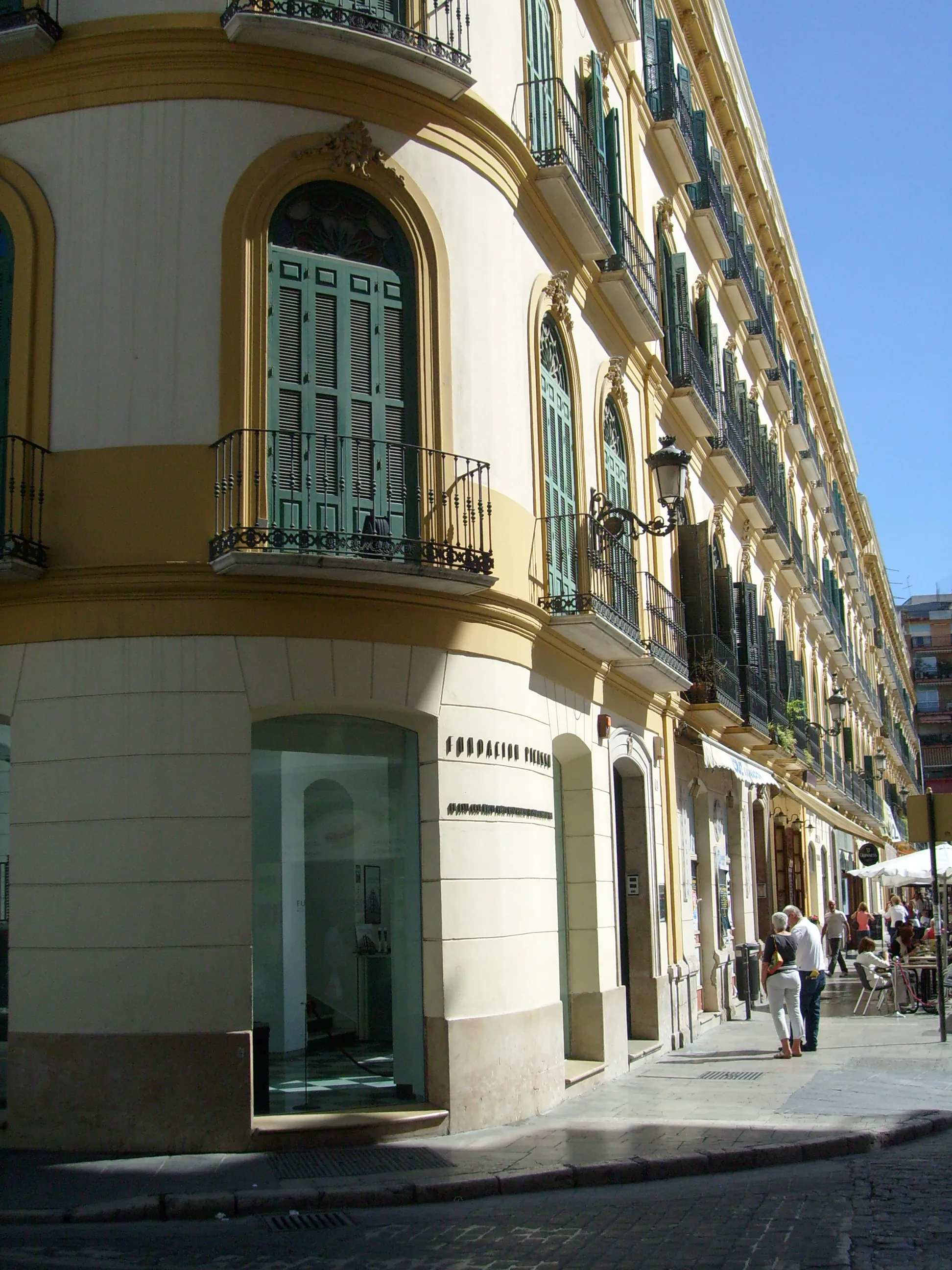 Vista de esquina del edificio amarillo y blanco del Museo Casa Natal Picasso con ventanas arqueadas, contraventanas verdes, balcones de hierro y peatones en la acera
