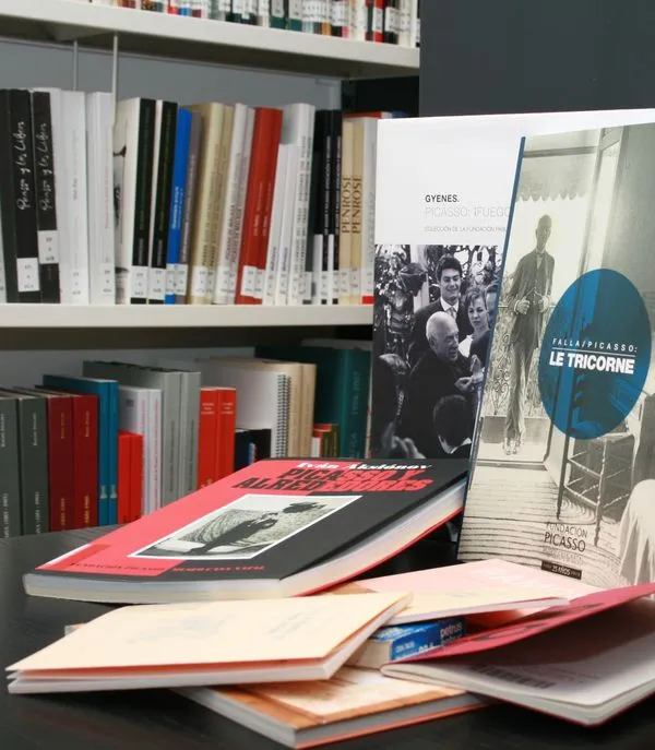 Libros y publicaciones sobre Pablo Picasso dispuestos sobre una mesa con estanterías al fondo, incluyendo un libro titulado Le Tricorne y un gran volumen de tapa roja.