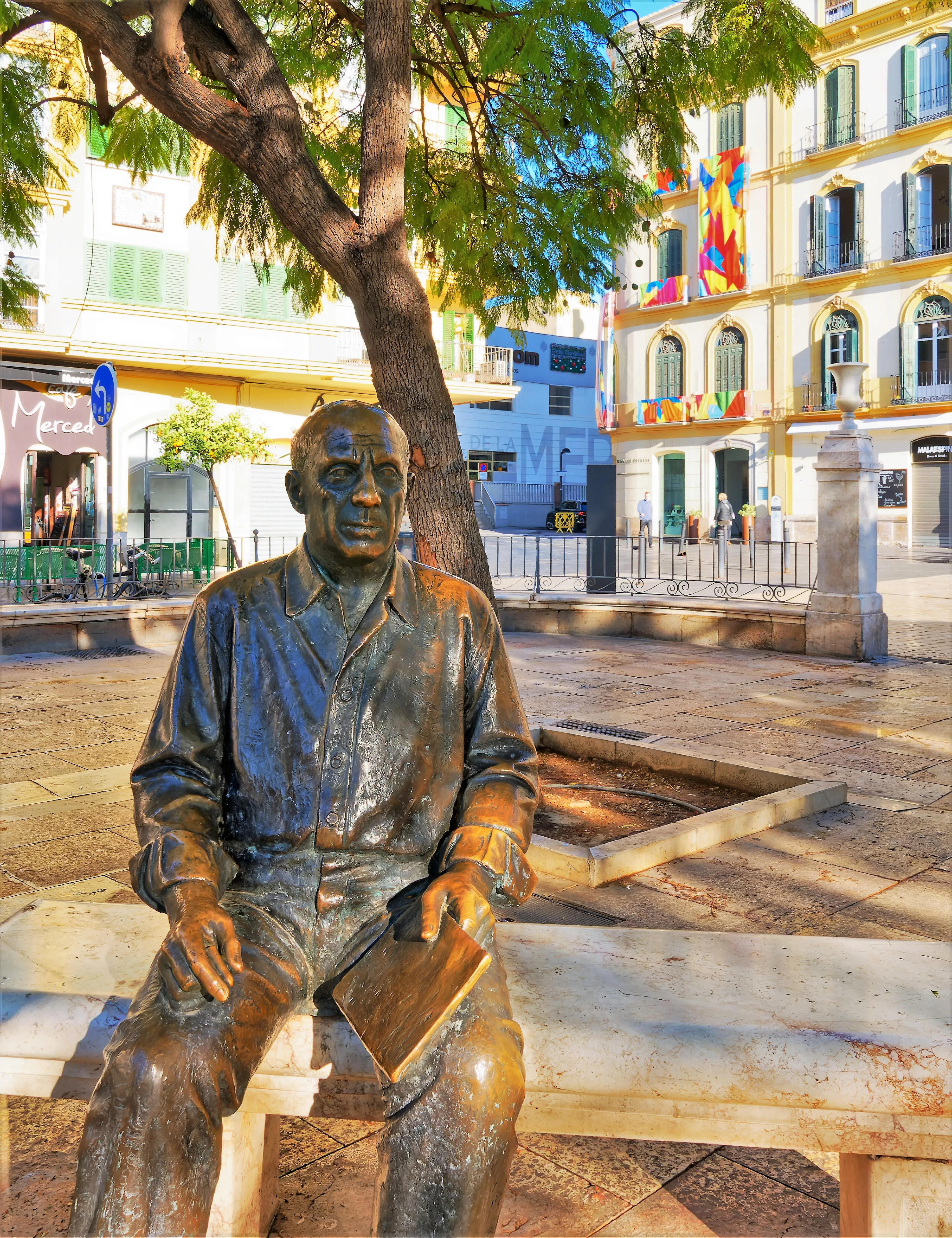 Estatua de bronce de Pablo Picasso sentado en un banco de piedra en la Plaza de la Merced, con el edificio del Museo Casa Natal Picasso y coloridas banderas visibles al fondo.