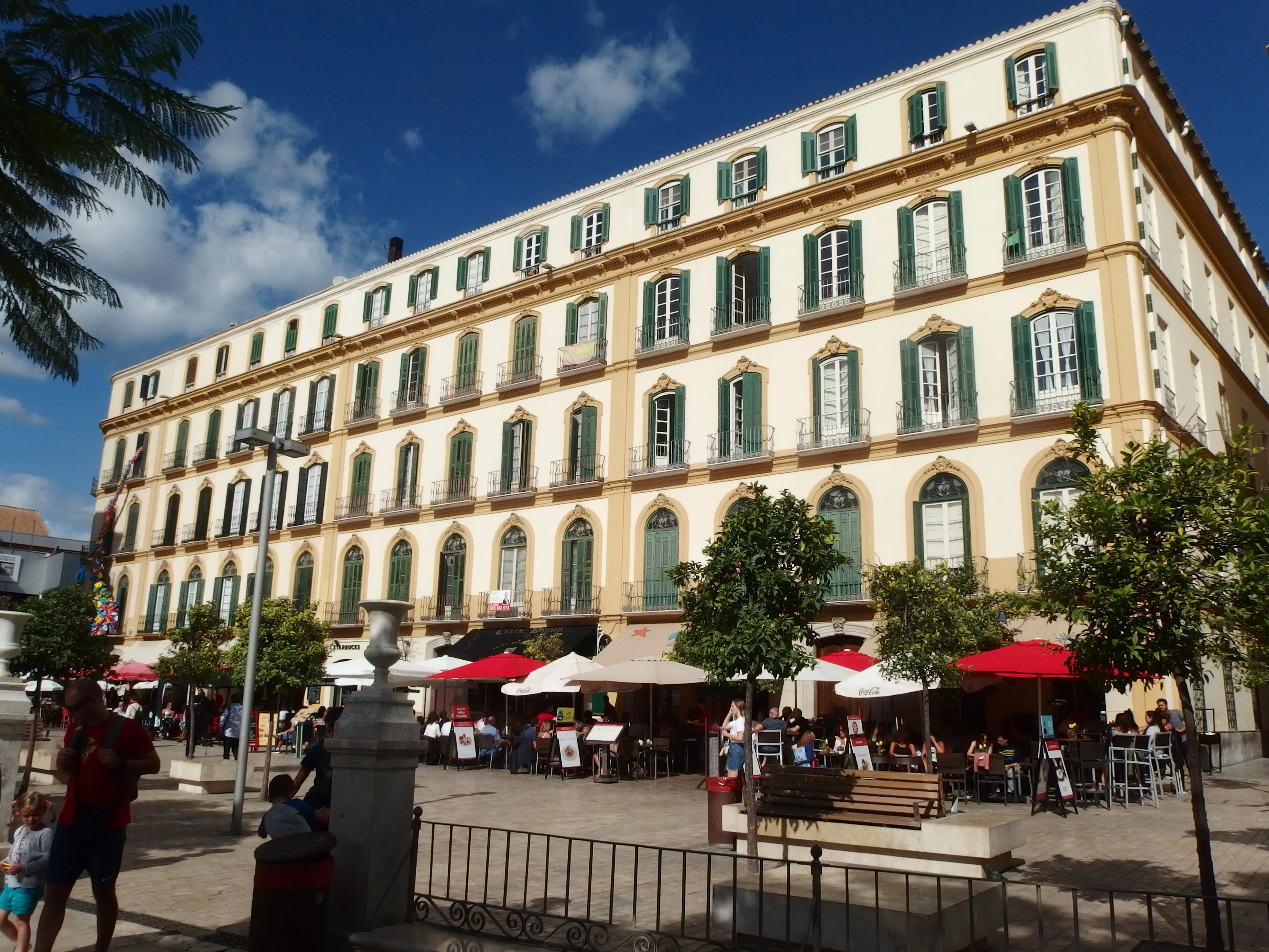 Gran edificio amarillo con contraventanas verdes que da a una plaza con mesas de cafetería al aire libre y sombrillas rojas bajo un cielo azul