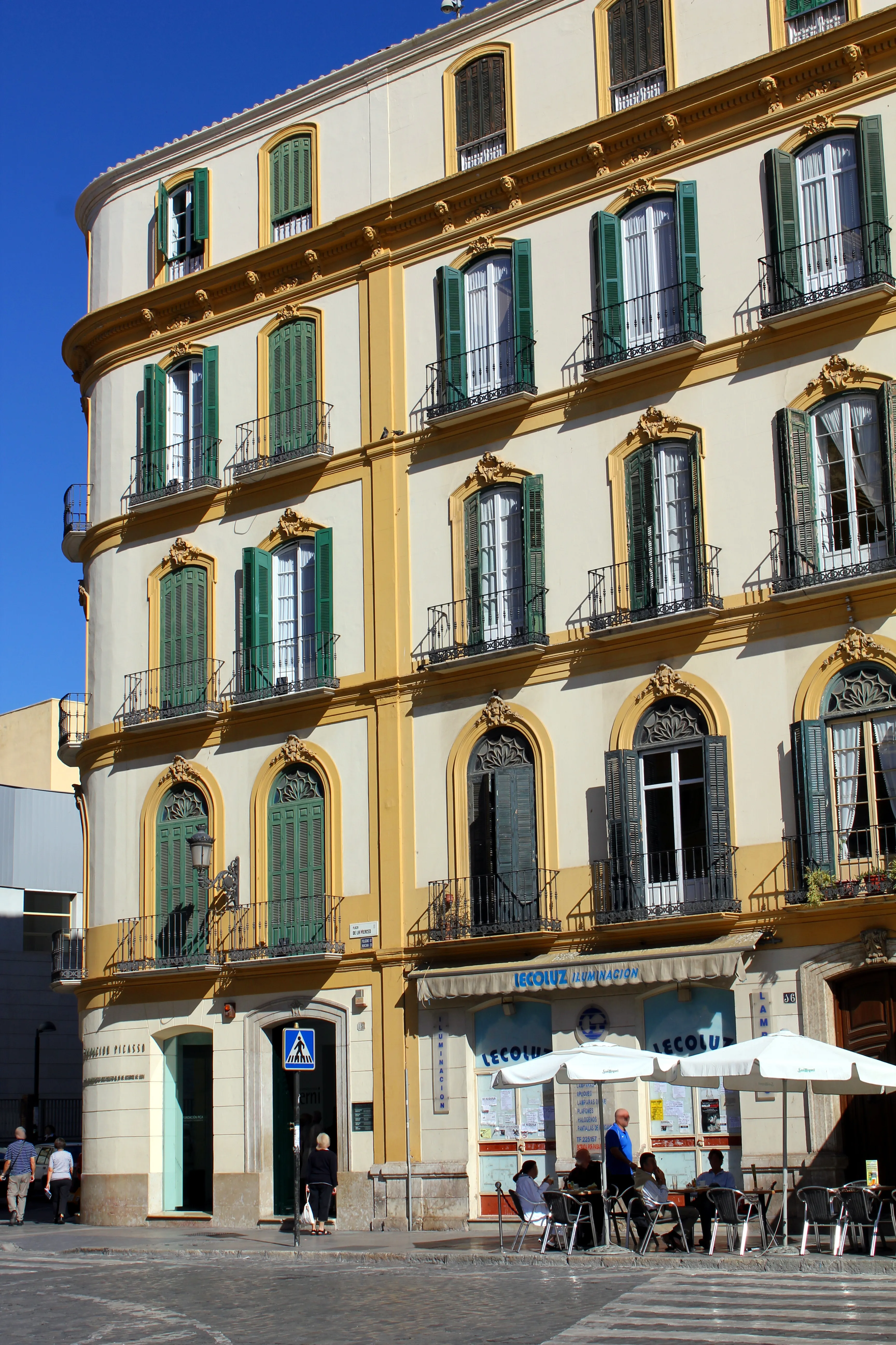 Edificio de varias plantas en tonos crema y amarillo con contraventanas verdes, ventanas arqueadas y balcones de hierro, lugar de nacimiento de Pablo Picasso en Málaga, con una cafetería y terraza en la planta baja