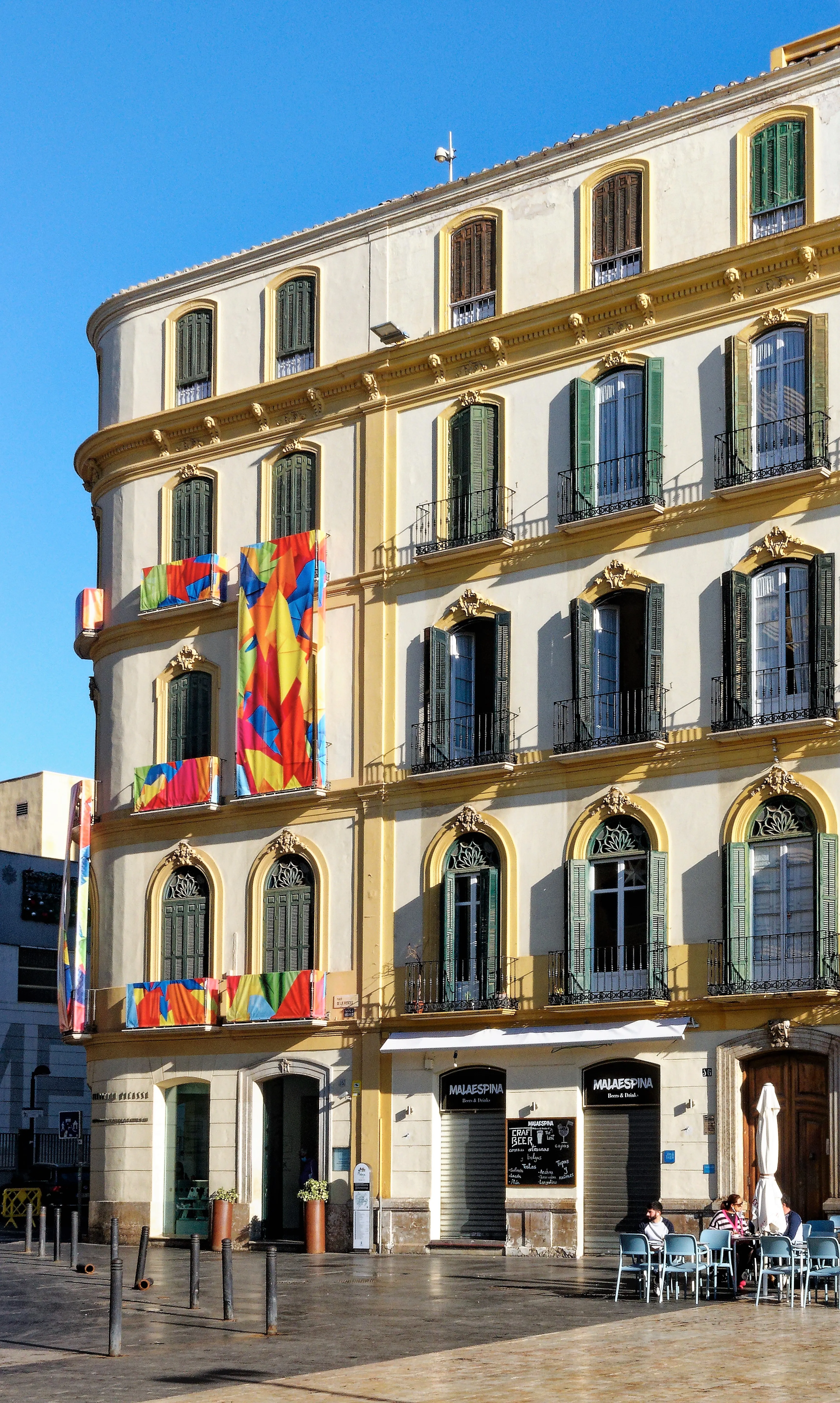 Fachada exterior del Museo Casa Natal Picasso con paredes amarillas y blancas, contraventanas verdes, ventanas arqueadas y banderas coloridas que cuelgan de los balcones