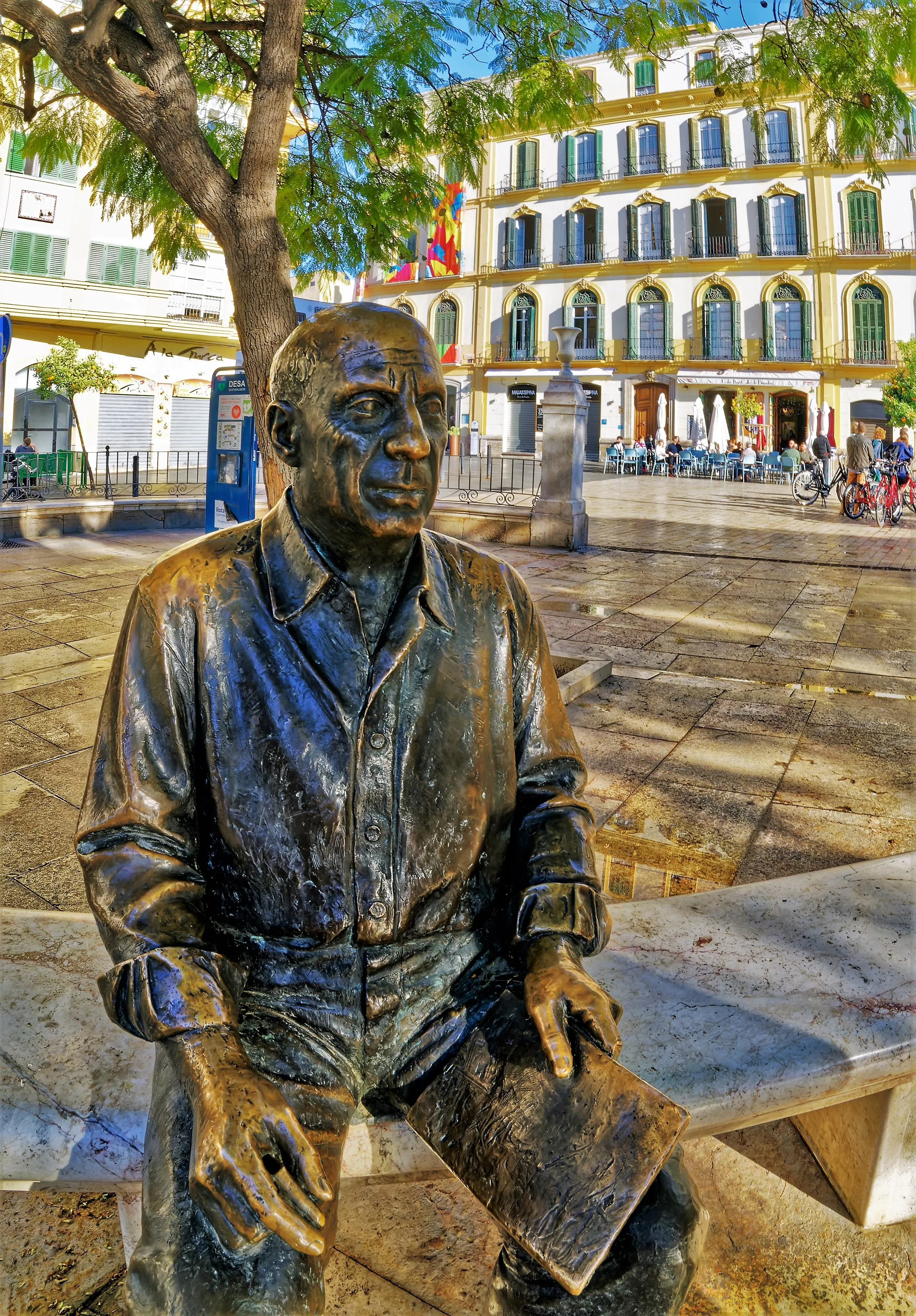 Estatua de bronce de Pablo Picasso sentado en un banco sosteniendo un cuaderno de bocetos en la Plaza de la Merced, Málaga, con edificios históricos y árboles al fondo