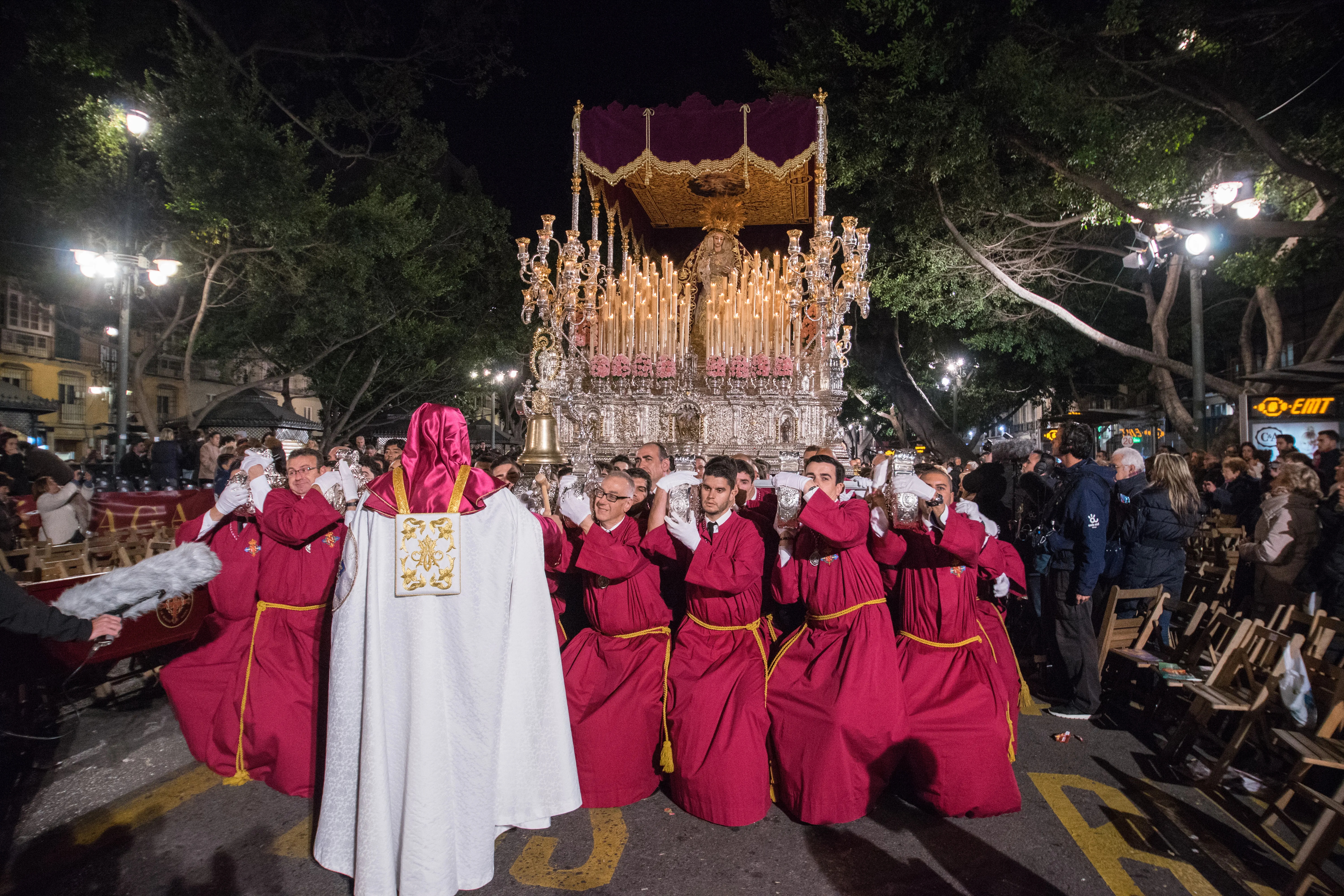 Night street procession with robed bearers in purple and white garments carrying a large ornate gilded religious float under streetlights