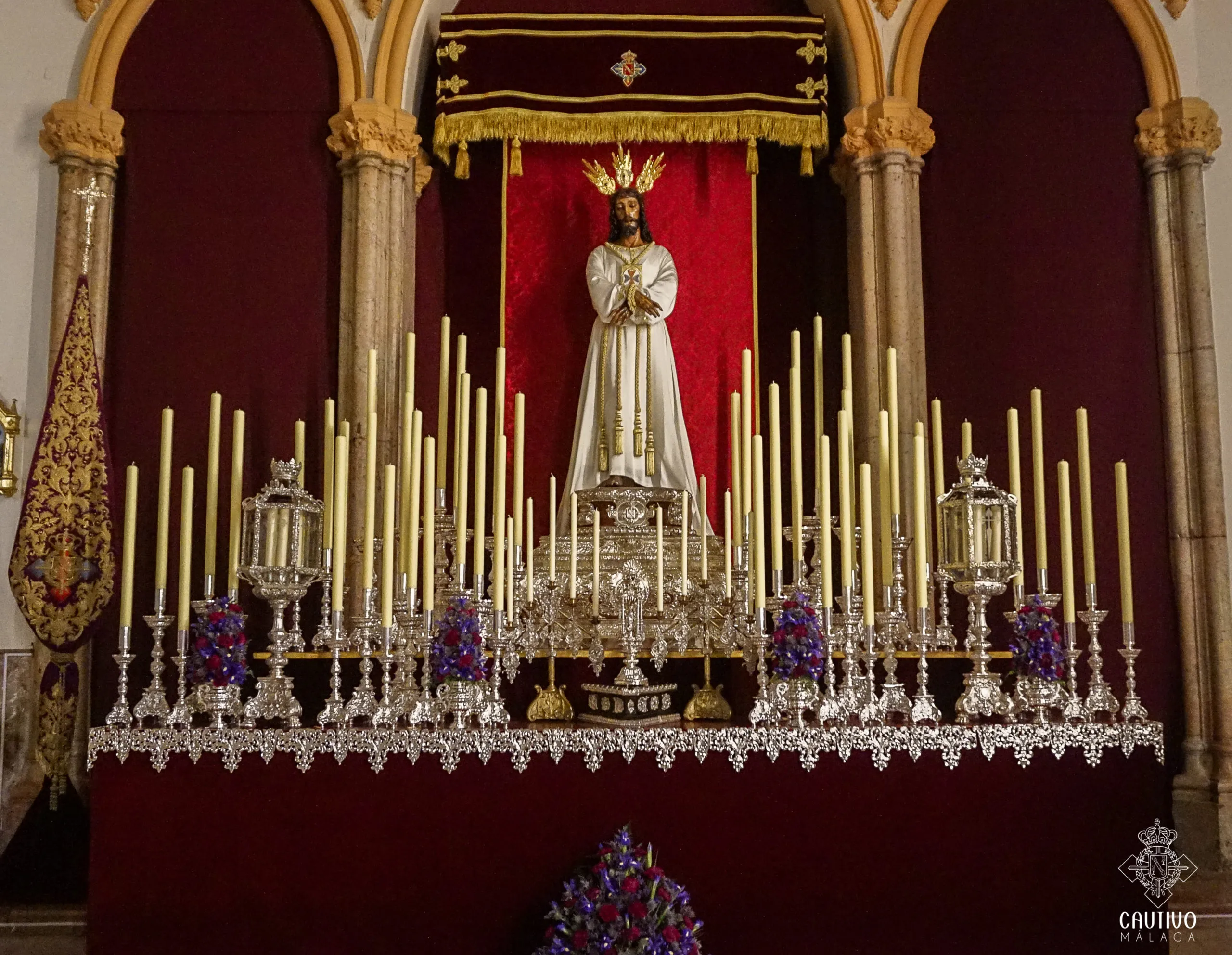 A processional throne featuring a white-robed statue of Jesus wearing a crown of thorns, surrounded by dozens of tall white candles, ornate silver lanterns, and purple floral arrangements against a deep red velvet backdrop framed by gold-trimmed arches