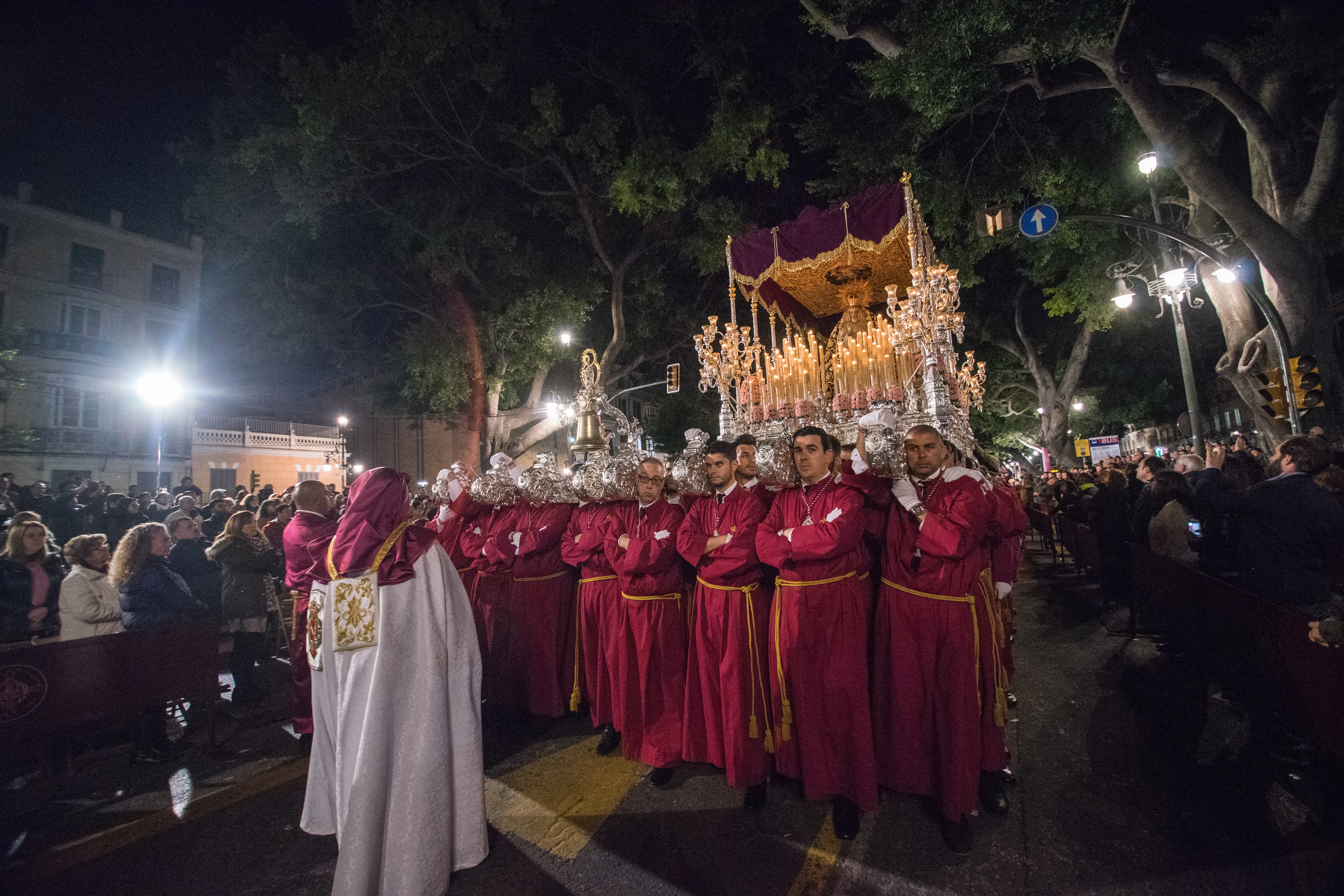 Procesión religiosa nocturna con portadores vestidos con túnicas color burdeos que llevan un gran trono procesional adornado y con velas a través de una calle arbolada llena de espectadores.