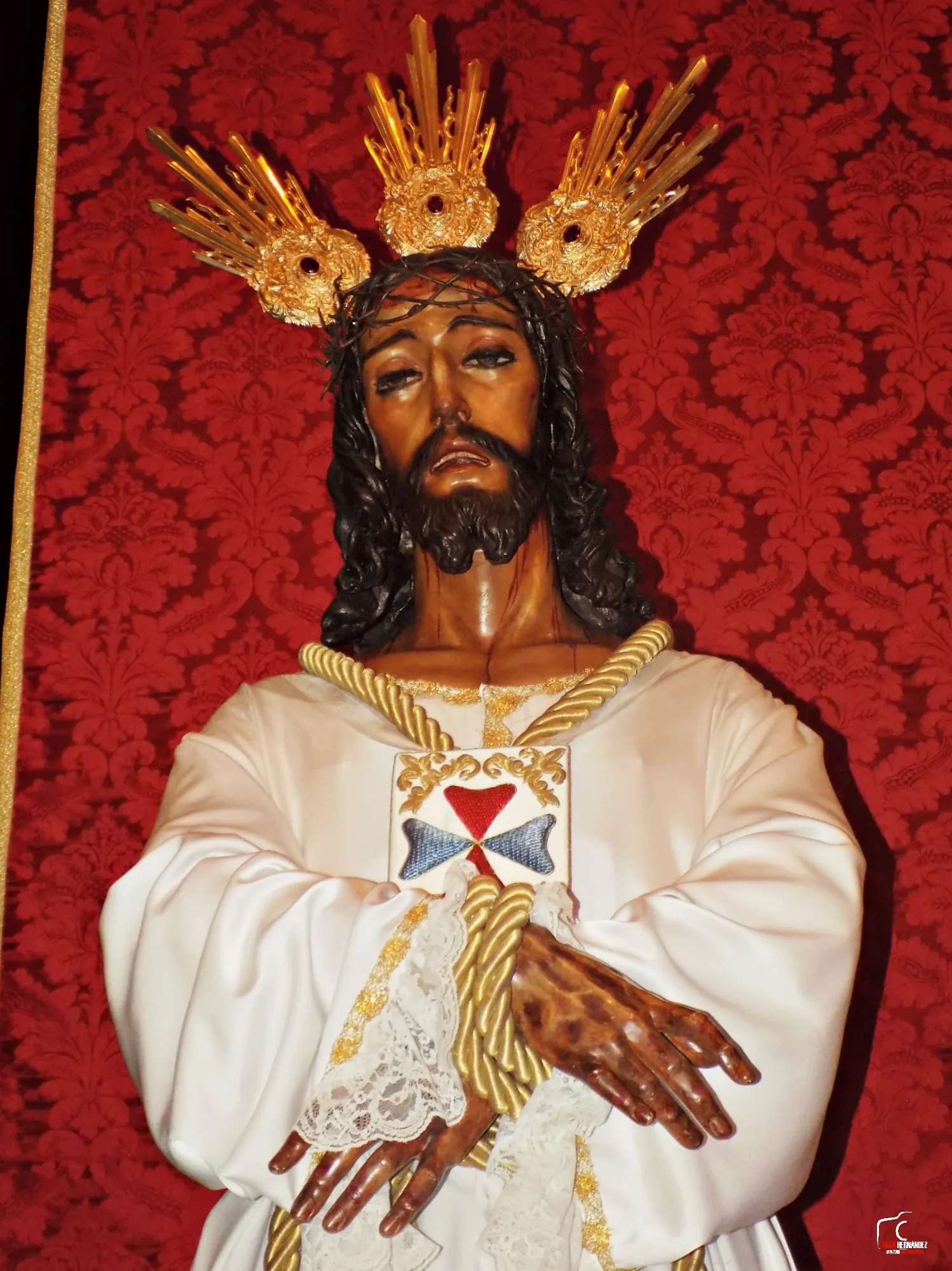 Estatua de Jesús Cautivo con corona de espinas y potencias doradas, vestido con túnica blanca con cíngulo y emblema de cruz, sobre un fondo con estampado rojo.