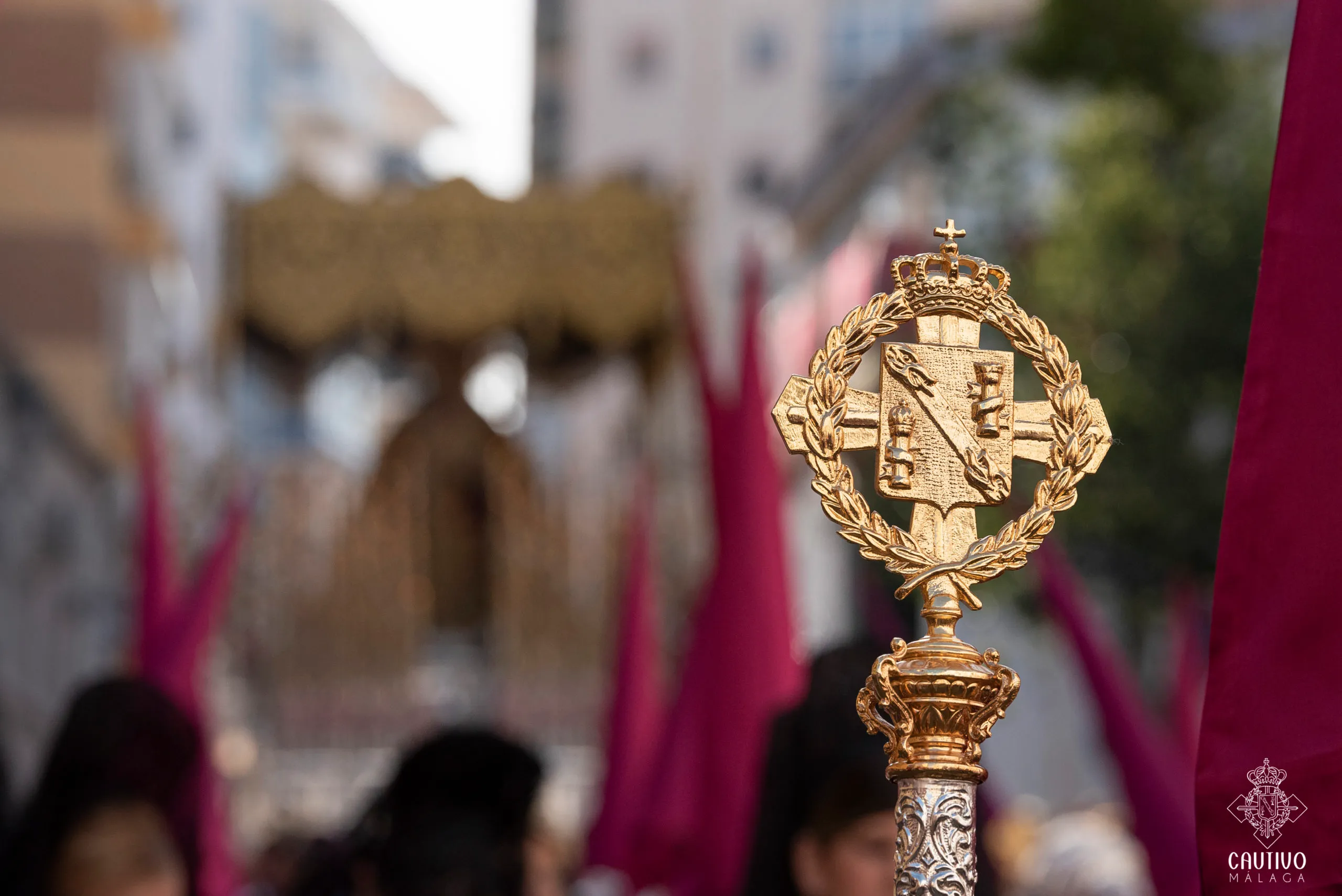 Báculo ceremonial dorado rematado con un escudo de armas coronado y un emblema de cruz, con figuras difusas vestidas con túnicas moradas y una carroza procesional al fondo.