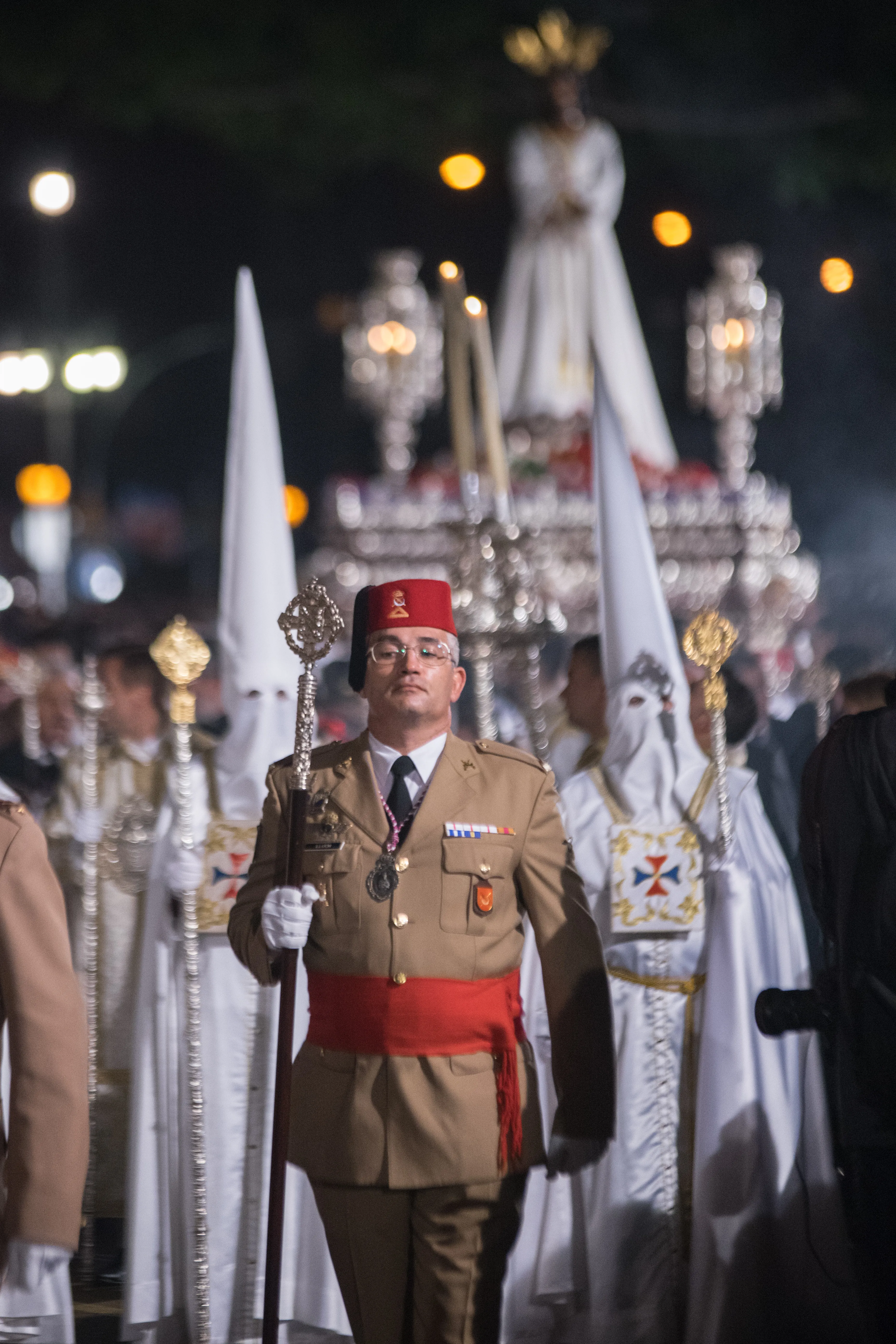 A man in a tan military-style uniform with a red fez and red sash holds a ceremonial staff during a nighttime procession, with white-robed nazarenos and a silver processional float carrying a statue of the Virgin Mary behind him.